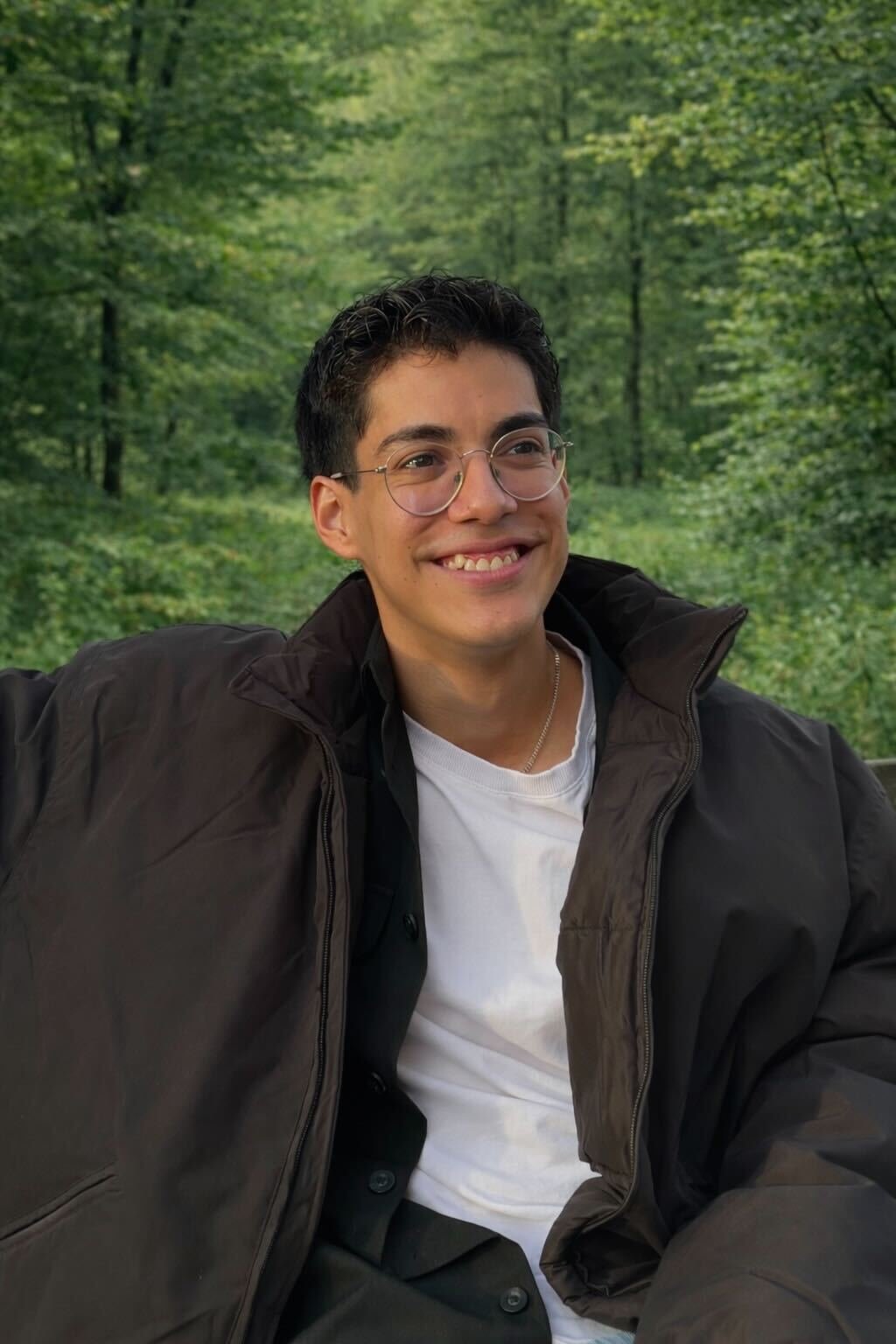 A young man with glasses and dark hair smiling outdoors in a green forest, wearing a black jacket over a white shirt.