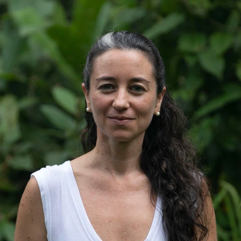 A woman with dark curly hair, wearing pearl earrings and a white sleeveless top, standing outdoors with green foliage in the background.