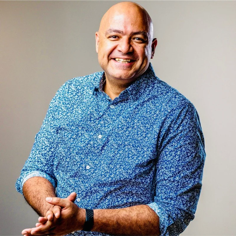 A smiling man with a bald head and light skin, wearing a blue floral button-up shirt, poses against a plain beige background.