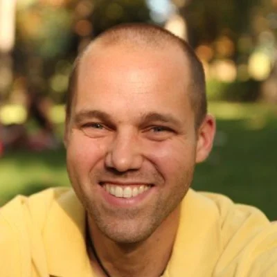 A smiling man with a shaved head wearing a yellow shirt outdoors in a park with trees and sunlight in the background.