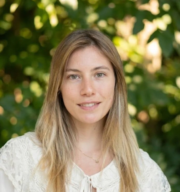A smiling young woman with long, light brown hair standing outdoors in a green, wooded area.
