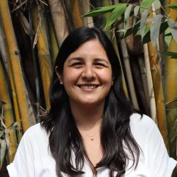 A young woman smiling outdoors in front of a bamboo fence and green plants.