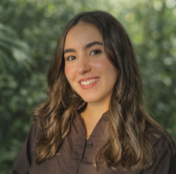 Young woman with long brown hair wearing a dark shirt, smiling in front of a lush green outdoor background.