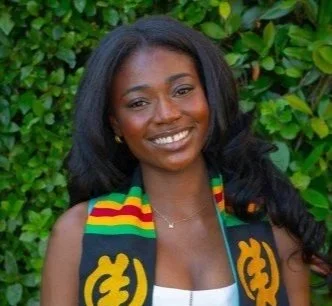 Young woman smiling outdoors, wearing a black scarf with colorful stripes and rasta symbols, and a white top.