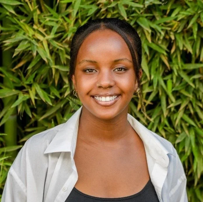 A young woman with dark skin and dark braided hair smiling in front of green leafy plants, wearing a white shirt over a black top.
