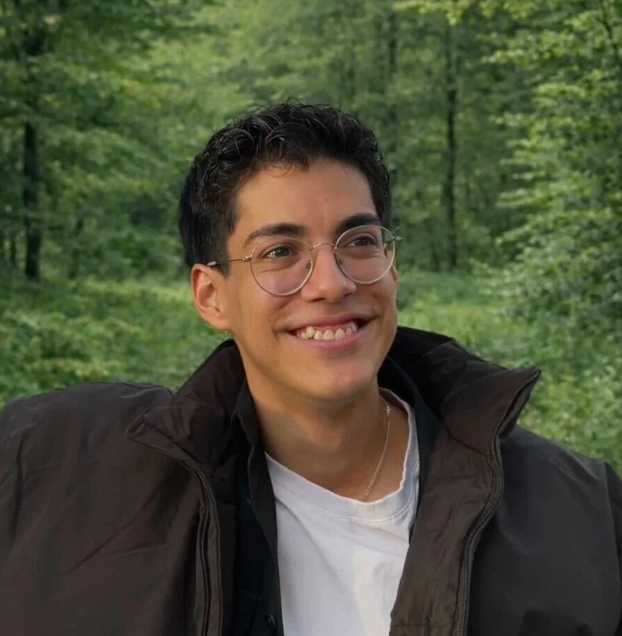 A young man with glasses and dark hair smiling outdoors in a green forest, wearing a black jacket over a white shirt.