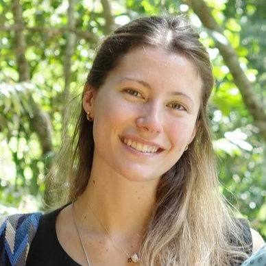 A smiling young woman with long, light brown hair standing outdoors in a green, wooded area.