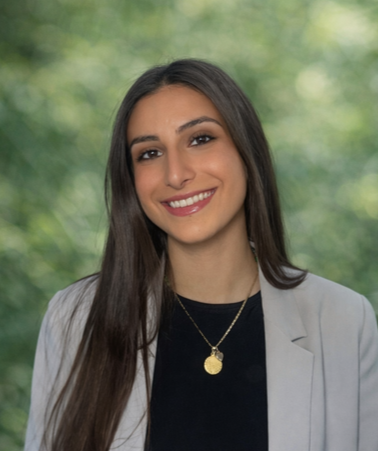 A young woman with long dark hair, smiling, wearing a gray blazer over a black top, with a gold necklace, standing outdoors with blurred green trees in the background.