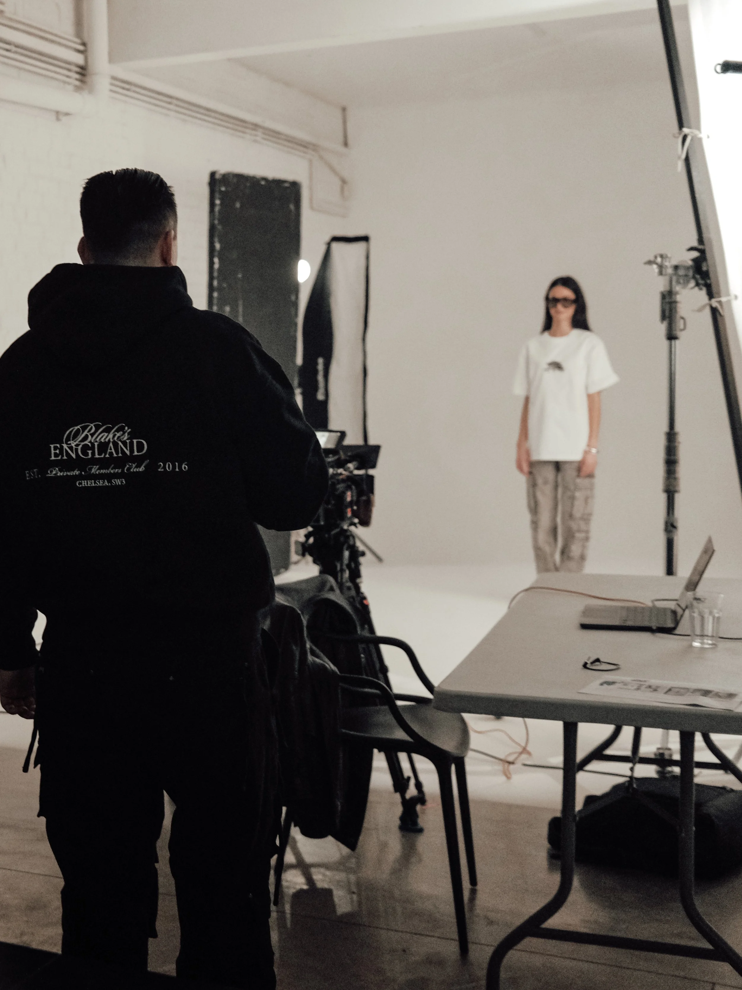 A photoshoot studio with a model standing in front of a white backdrop, wearing glasses, a white t-shirt, and patterned pants, while a photographer or crew member in black attire looks on.