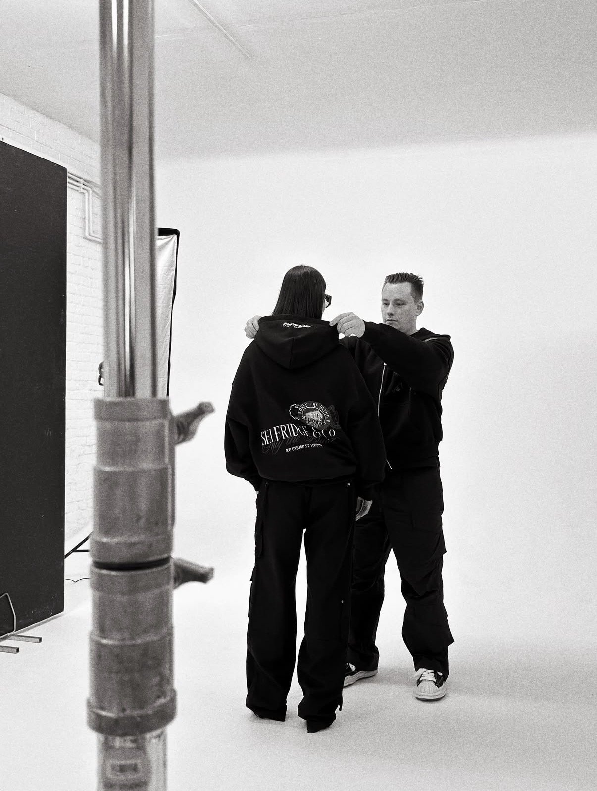 A man adjusting the hood of a woman in a photography studio, with a large reflector and white background.