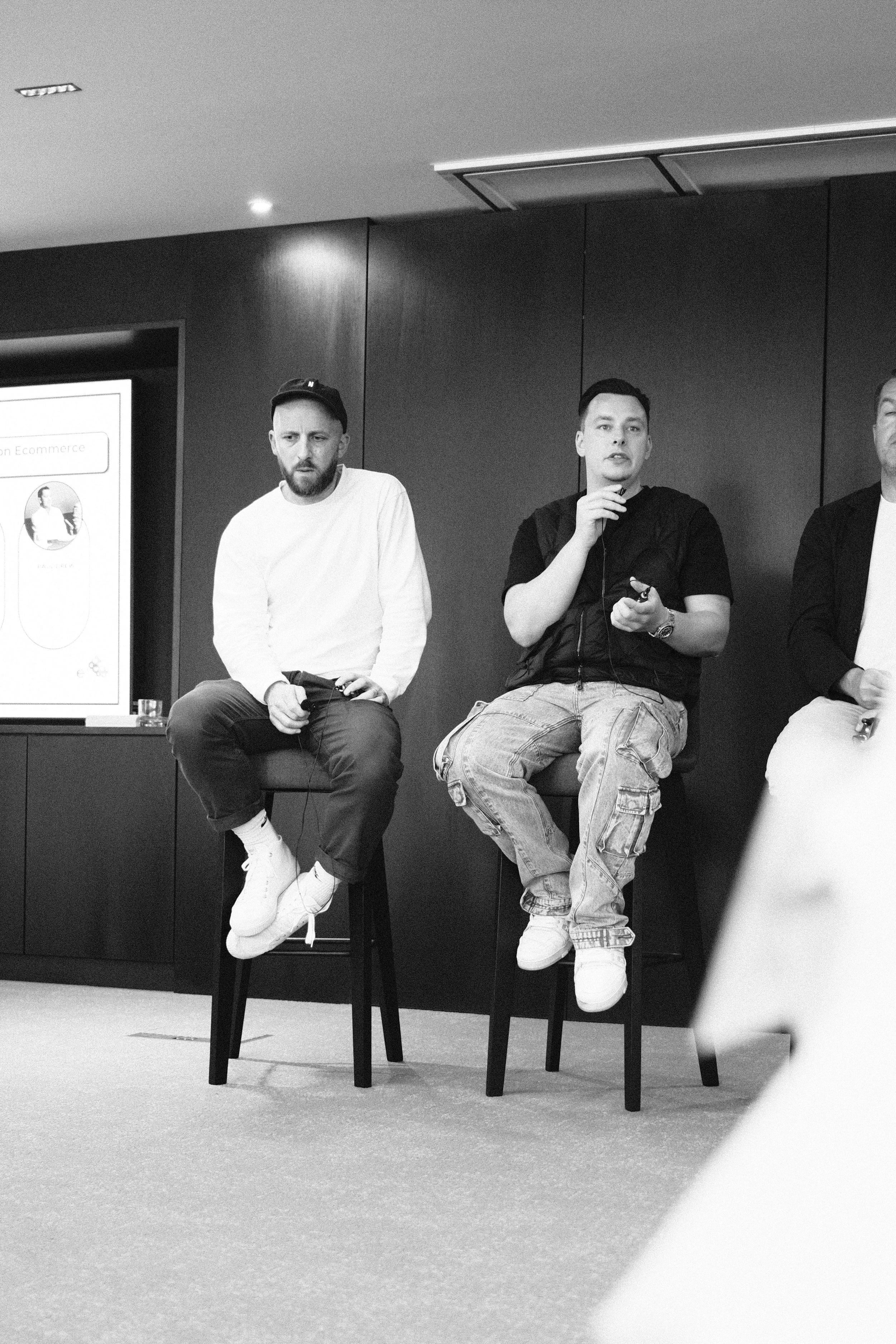 Three men sitting on high stools during a panel discussion, one speaking into a microphone, in a room with a presentation screen in the background.