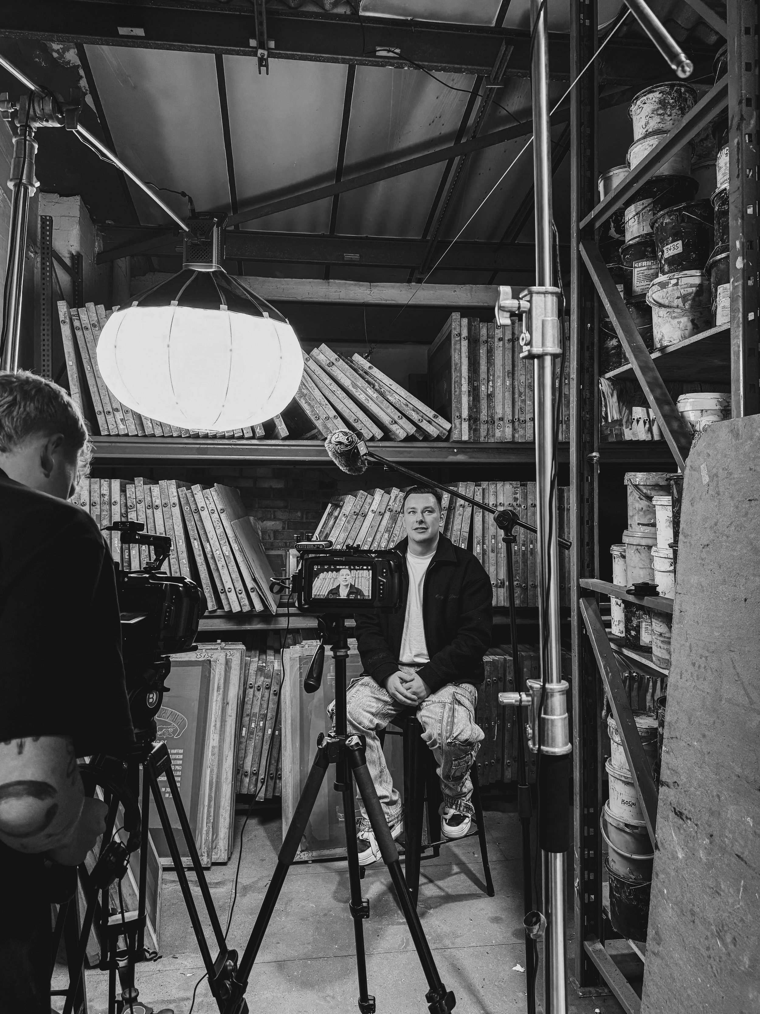 A man is sitting on a stool in front of a camera, filming himself during an interview or recording session in a cluttered workshop or storage space with shelves of cans and books behind him and a large glowing lamp overhead.