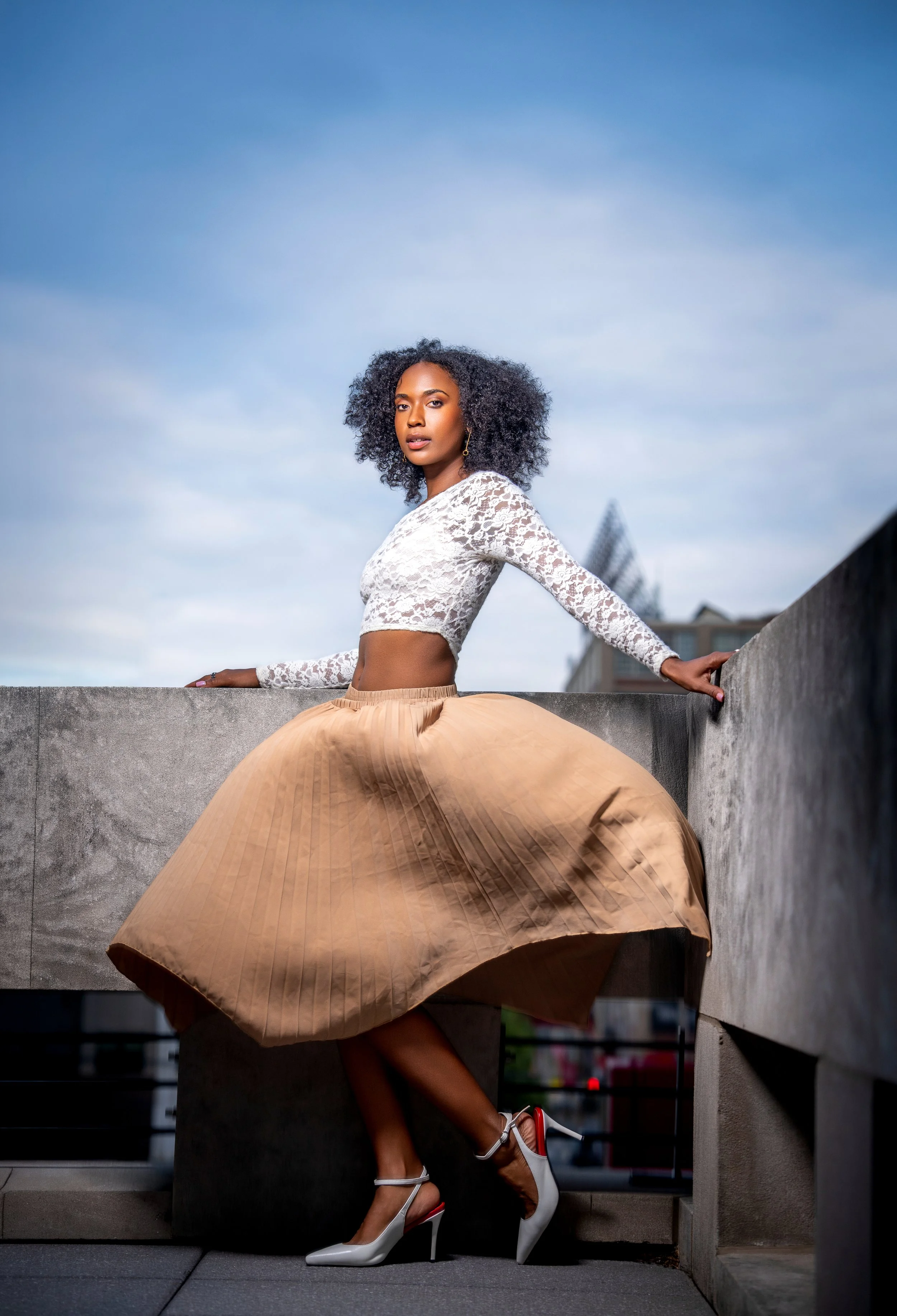 Editorial fashion portrait featuring a model in a flowing pleated skirt, photographed outdoors with dramatic movement, architectural lines, and soft blue skies.