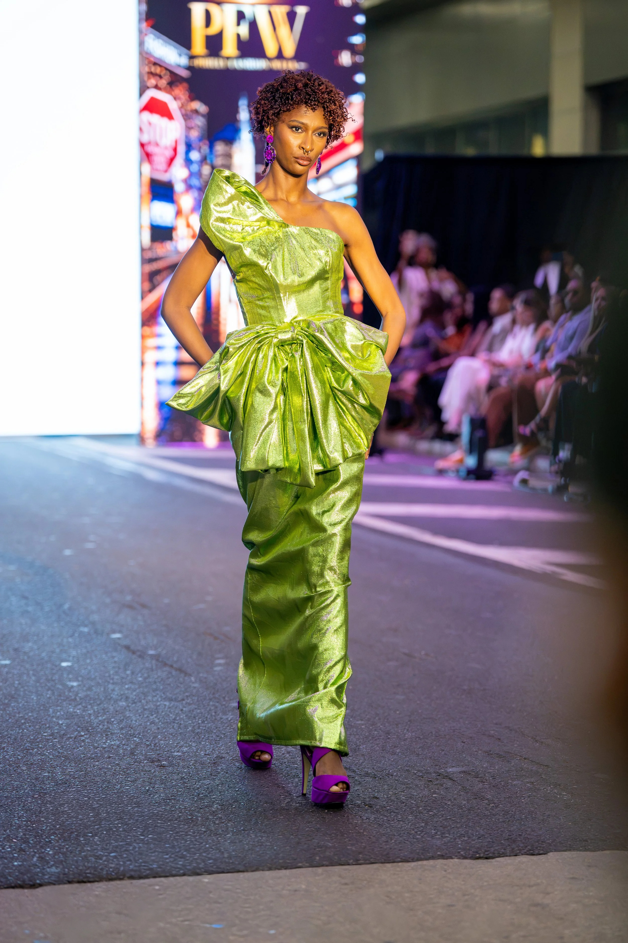 Model walking the Philly Fashion Week runway in a metallic lime green one-shoulder gown with an oversized sculptural waist detail and purple heels.