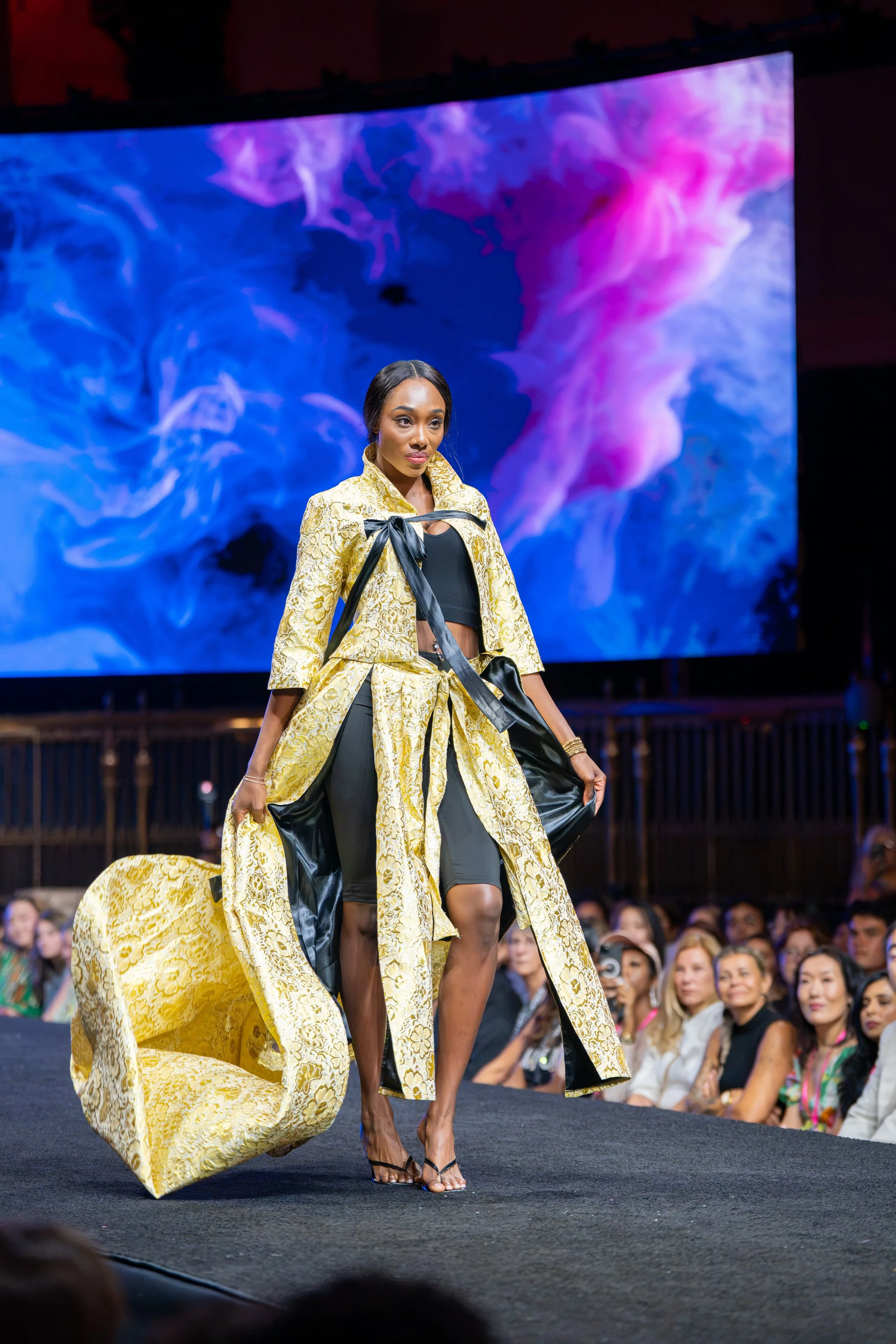 Model walking the New York Fashion Week runway in an elaborate gold brocade look with a dramatic sculptural trailing piece.
