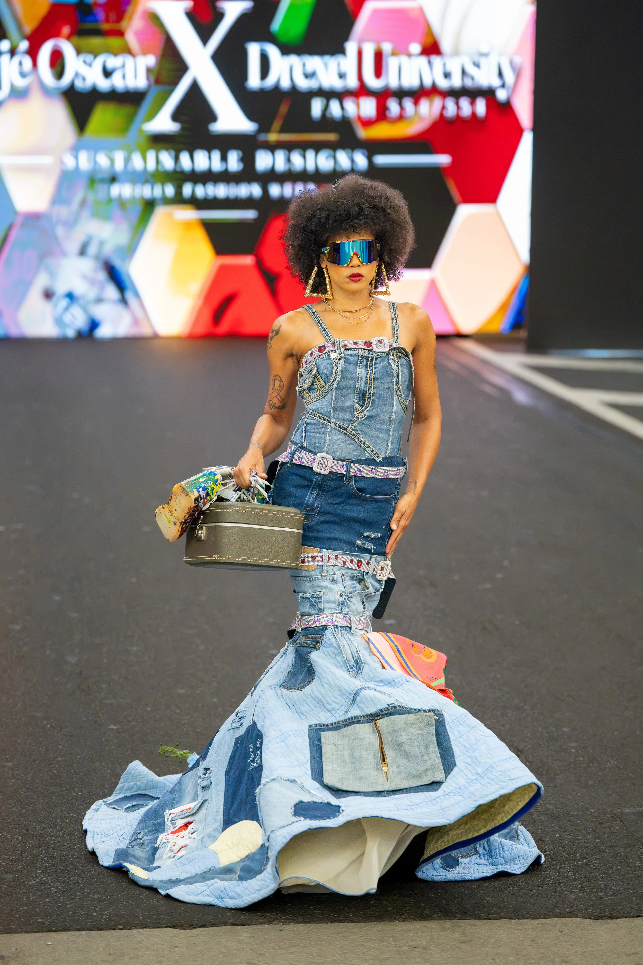 Model walking the Philly Fashion Week runway in an avant-garde reconstructed denim gown featuring patchwork details, oversized sunglasses, and statement accessories.