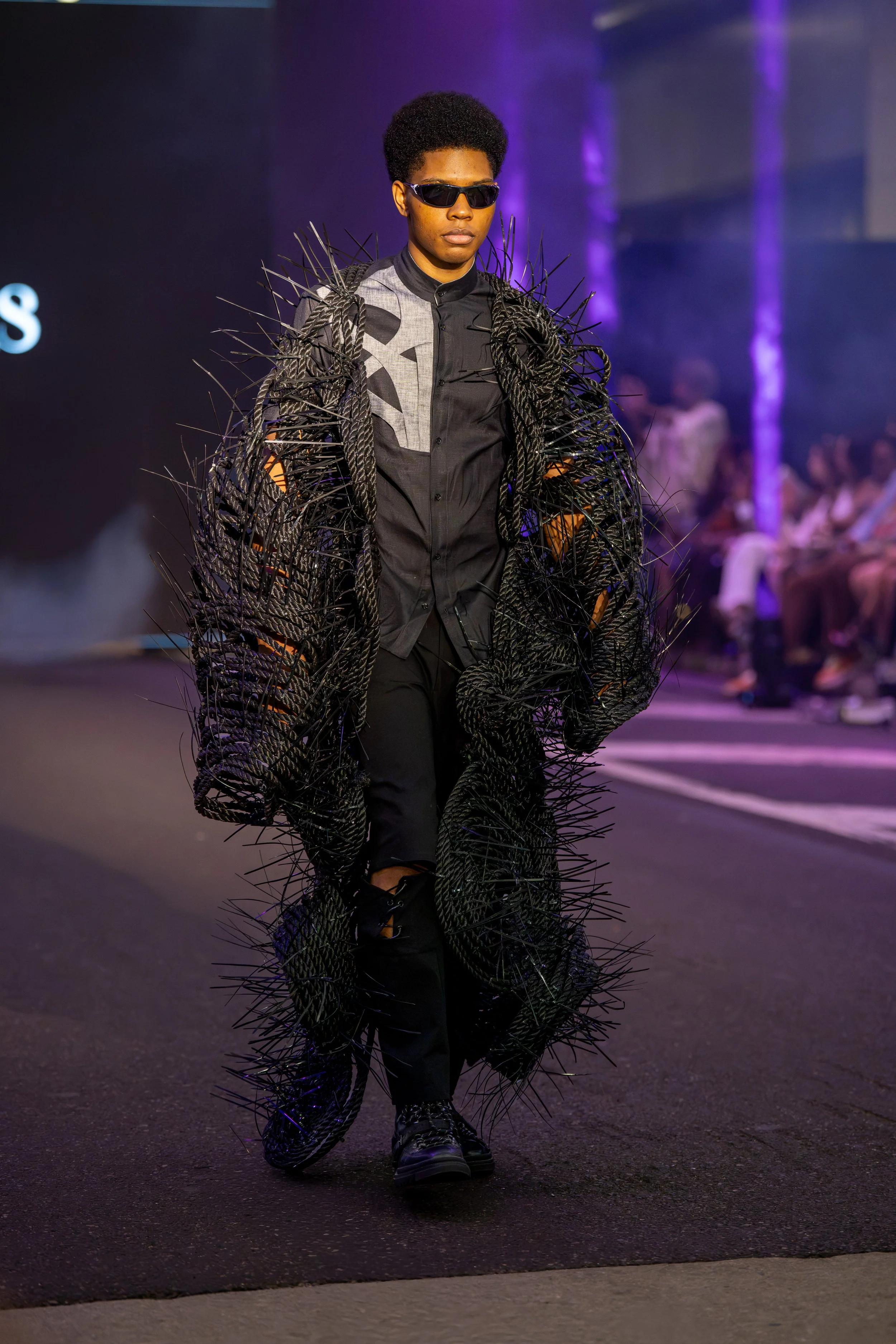 Model on the Philly Fashion Week runway wearing an experimental black sculptural outerwear design with intricate woven textures and dramatic three-dimensional detailing.