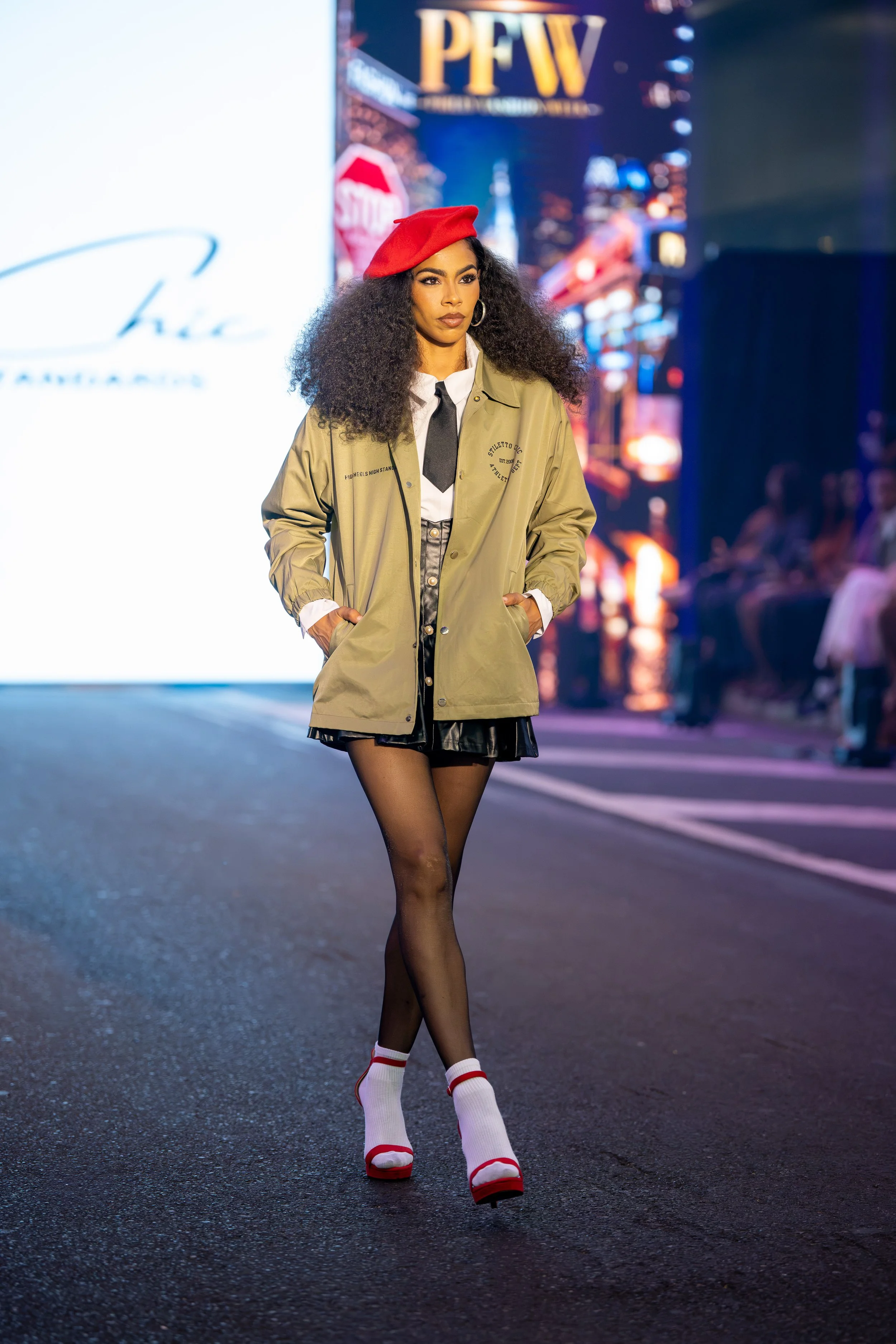 Model walking the Philly Fashion Week runway in a fashion-forward layered look featuring a red beret, khaki jacket, black mini skirt, tie, and statement heels.