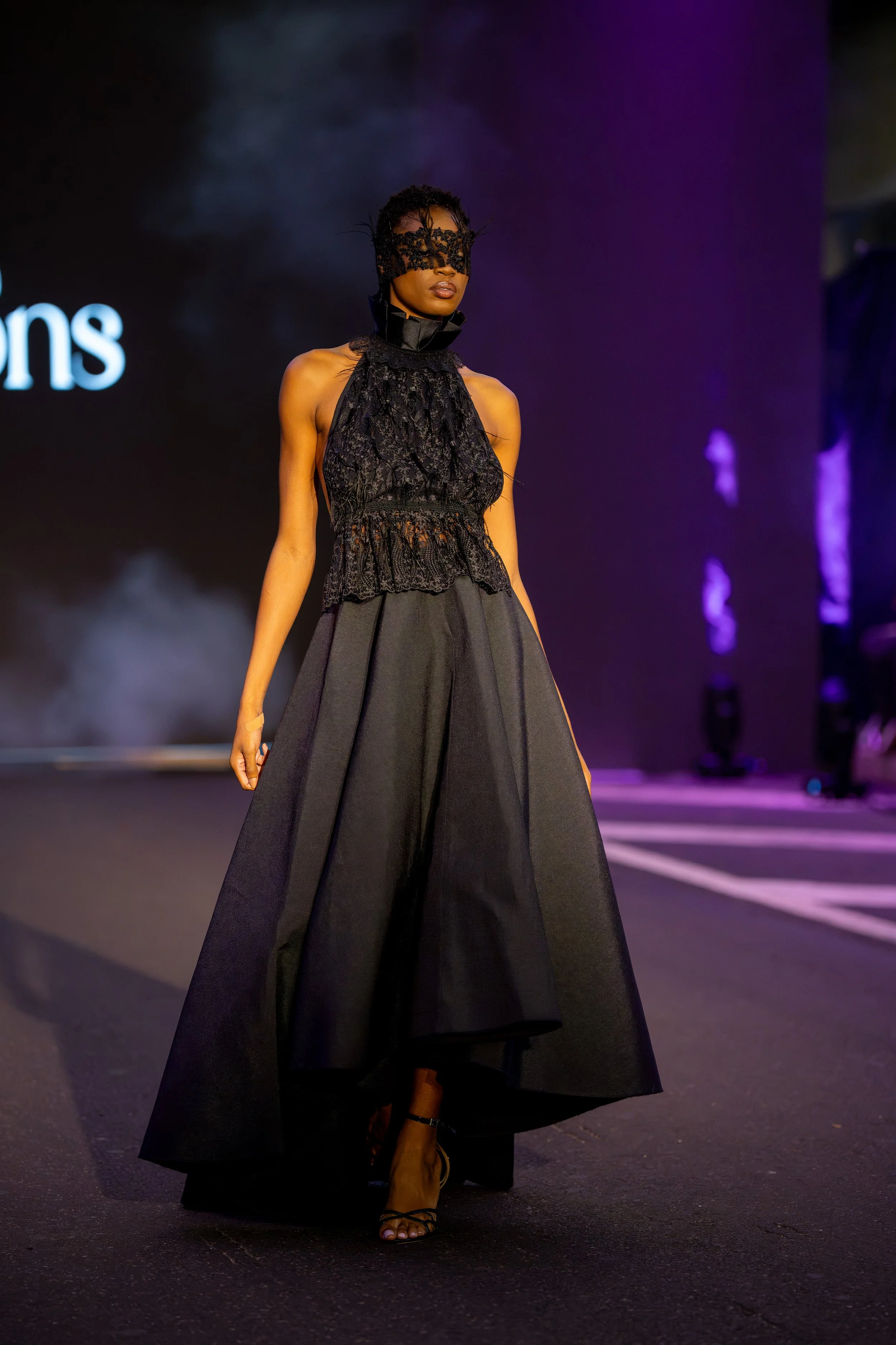 Model walking the Philly Fashion Week runway in an avant-garde black ensemble featuring a lace face covering, detailed halter bodice, and voluminous skirt.