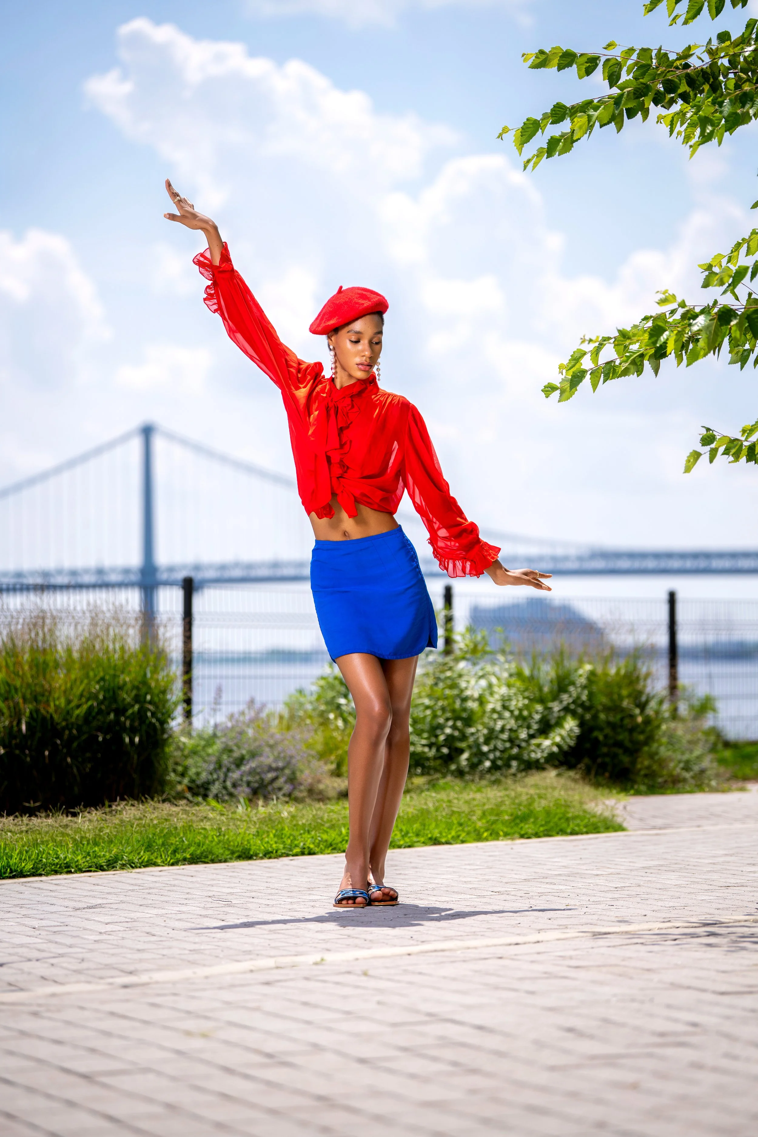 Outdoor editorial portrait of a fashion model posing gracefully in a vibrant red blouse, blue skirt and red beret with the Philadelphia waterfront and bridge in the background.