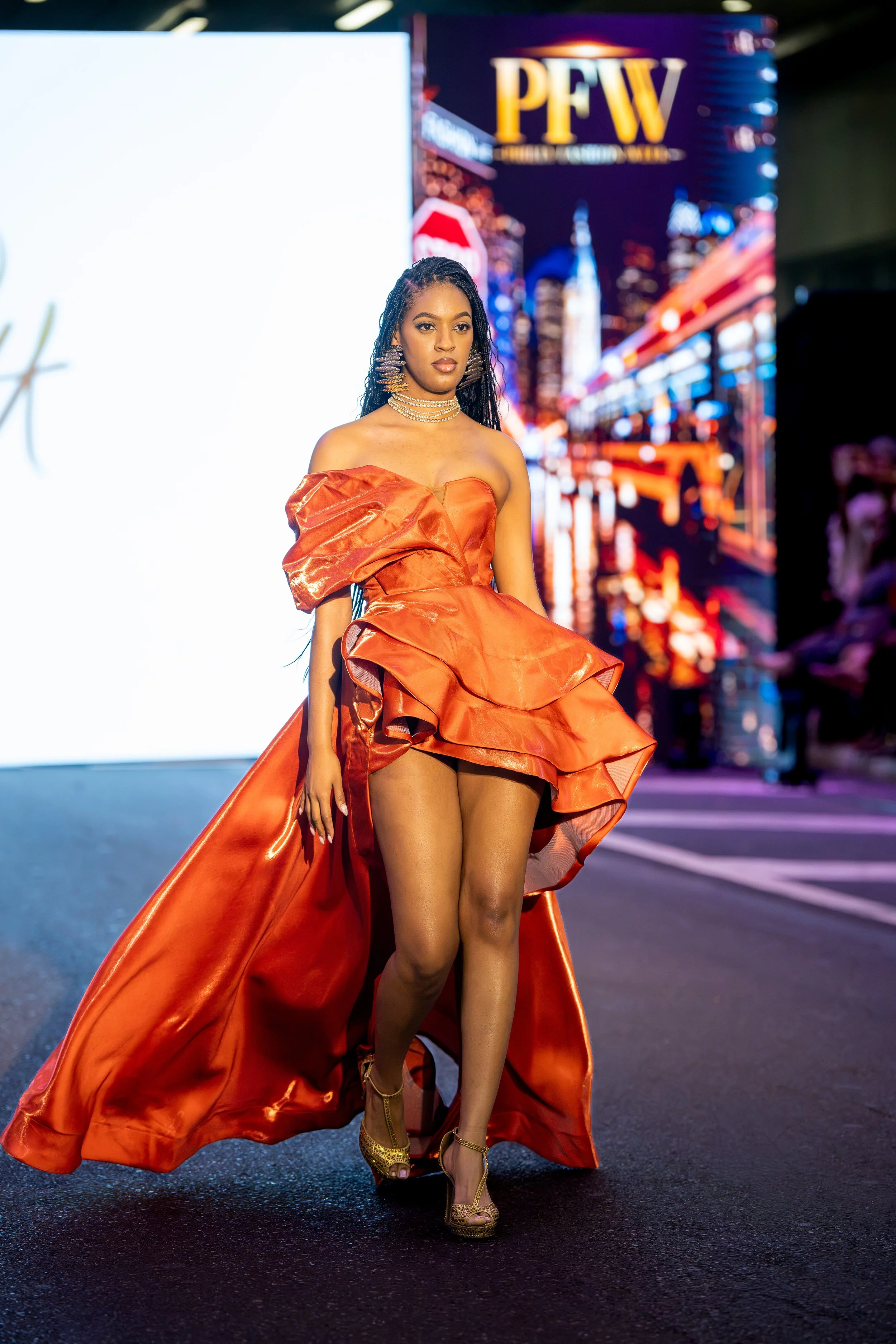 Model walking the Philly Fashion Week runway in a dramatic orange sculptural gown with an asymmetrical bodice, layered skirt, and sweeping train.