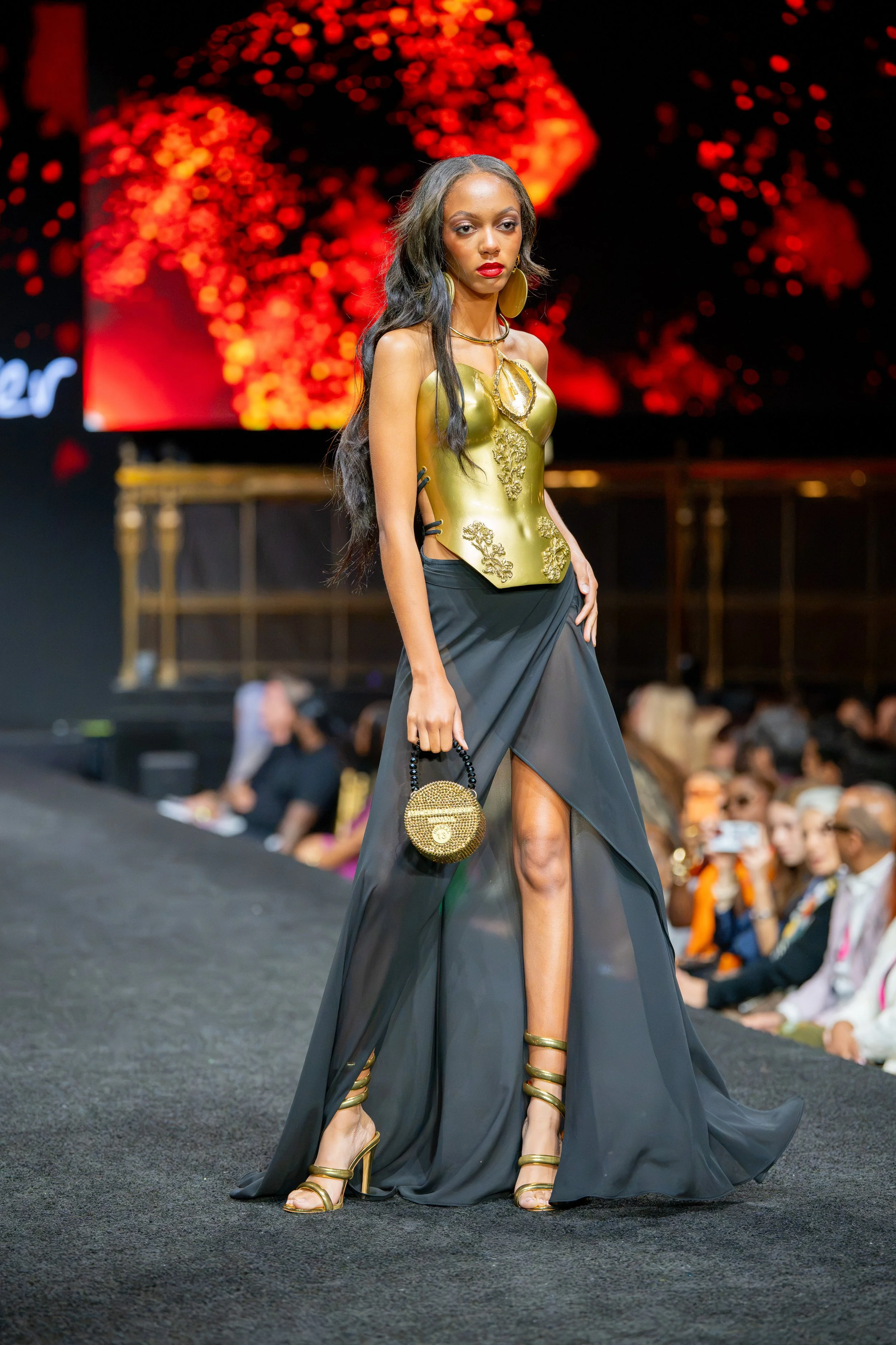 Model wearing a structured gold corset, long black skirt and round gold handbag on the New York Fashion Week runway at Gotham Hall.
