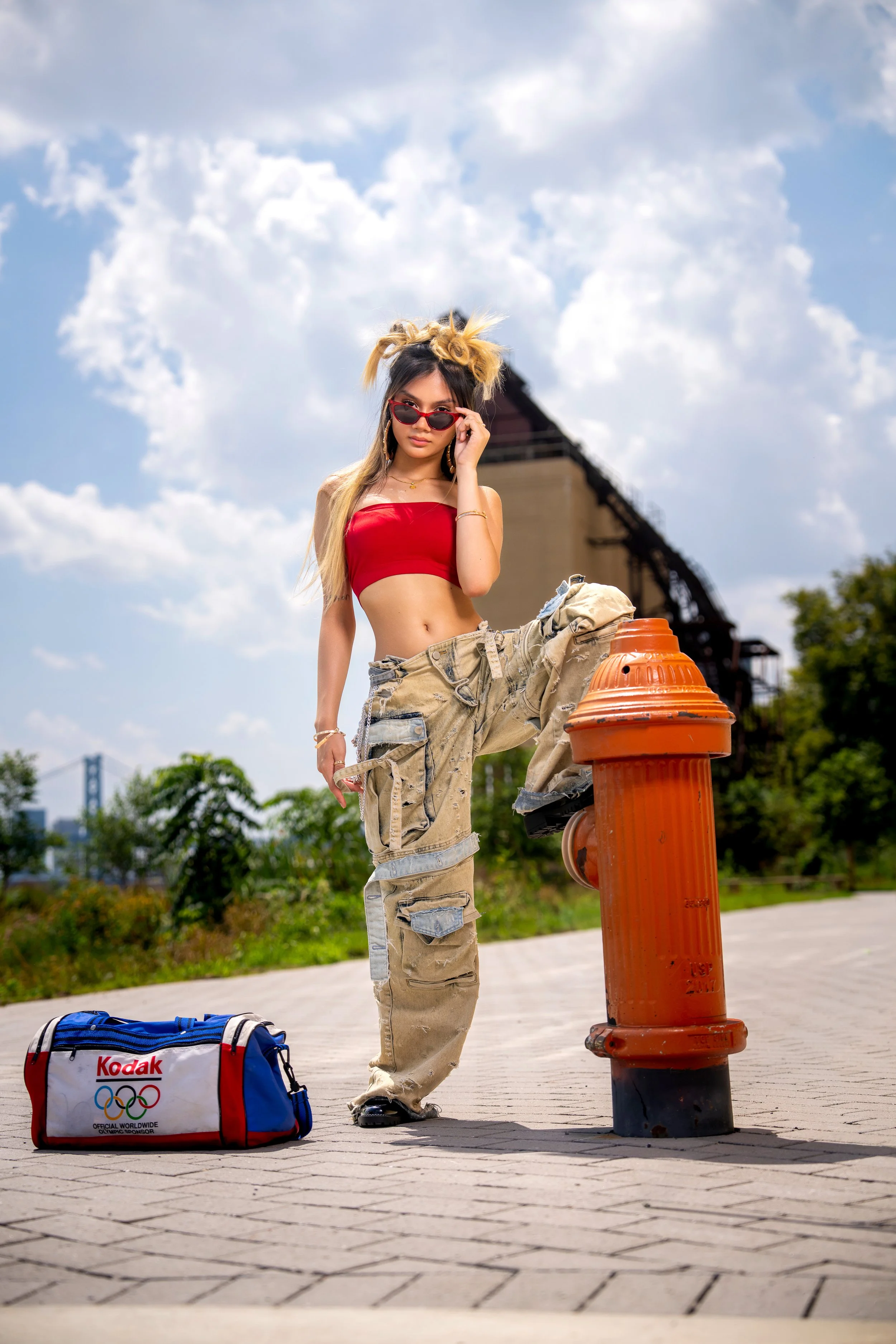Outdoor fashion portrait of a model posing beside an orange fire hydrant in distressed oversized pants, a red top and red sunglasses under a dramatic blue sky.