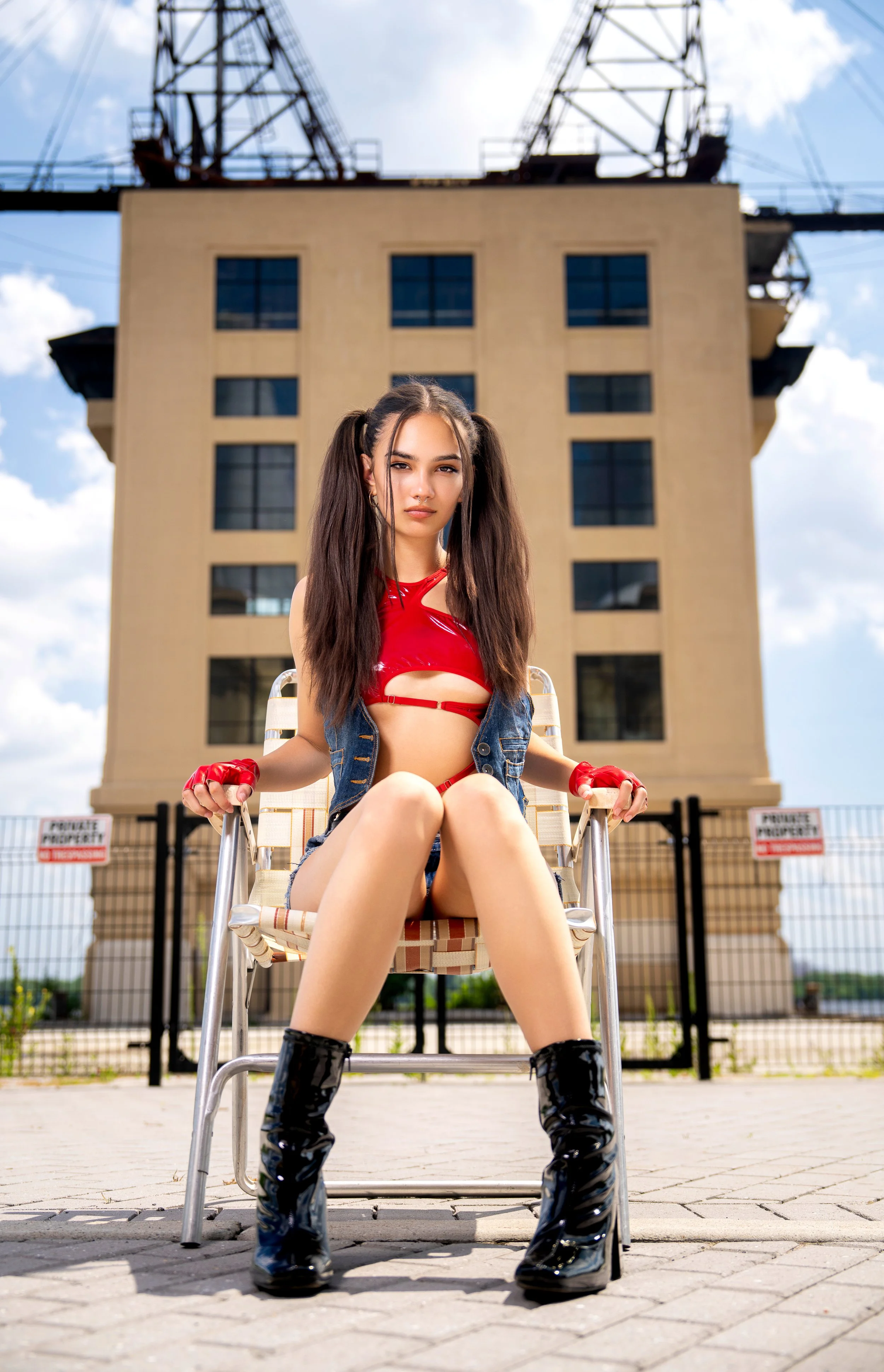 Editorial fashion portrait of a model seated confidently in a vintage chair wearing a red cutout top, denim vest, shorts and black platform boots.