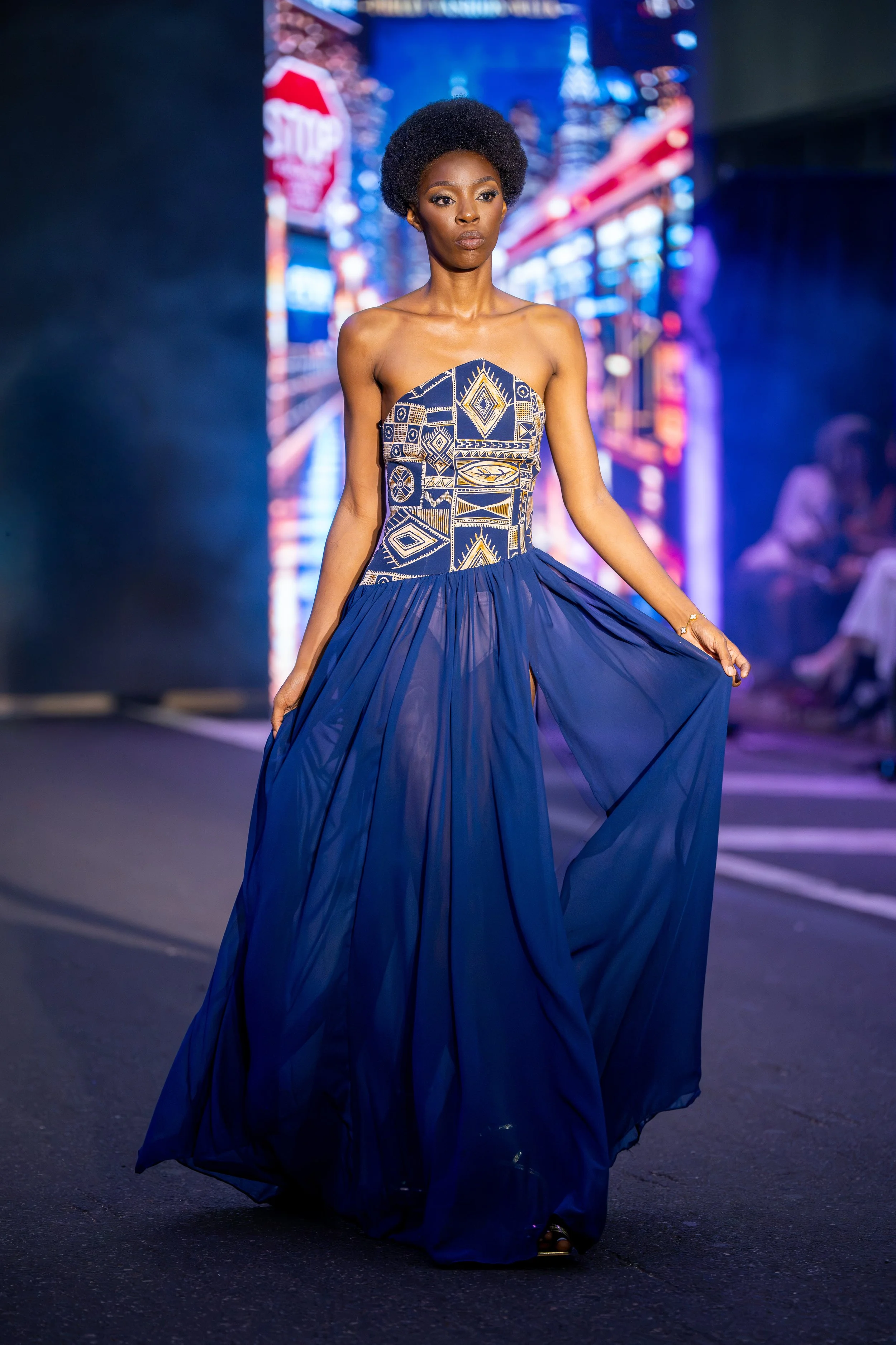 Model on the Philly Fashion Week runway wearing a flowing royal blue gown with a geometric patterned bodice and dramatic sheer skirt.