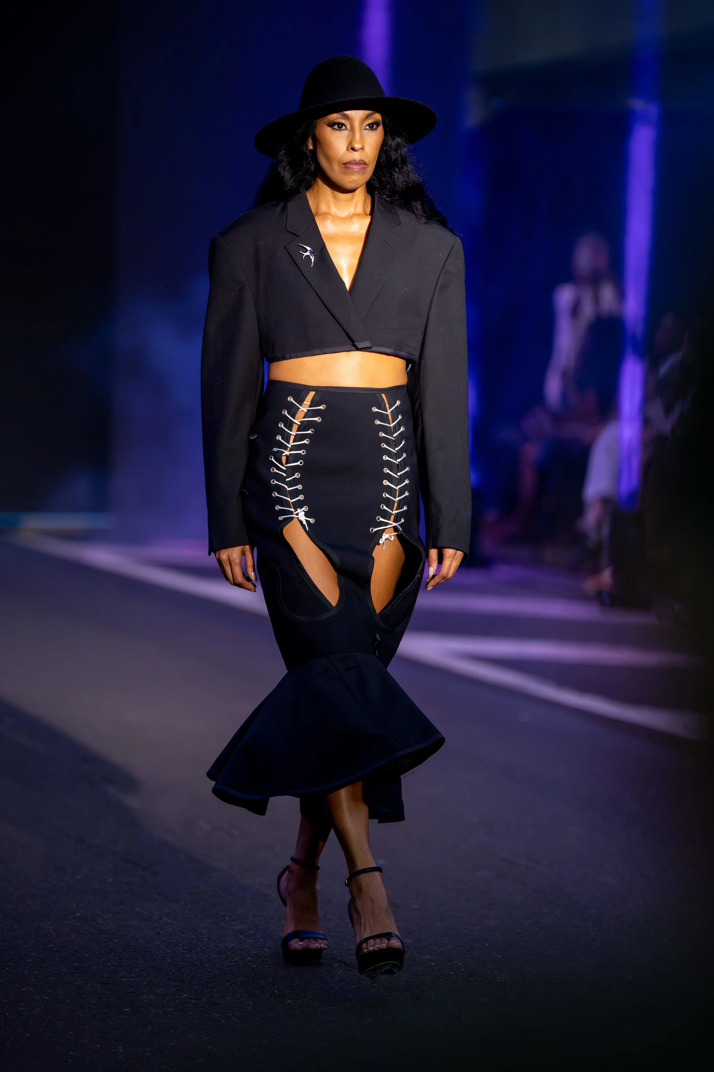 Model on the Philly Fashion Week runway wearing a black cropped blazer, wide-brim hat, and dramatic lace-up skirt with sculptural cutout detailing.