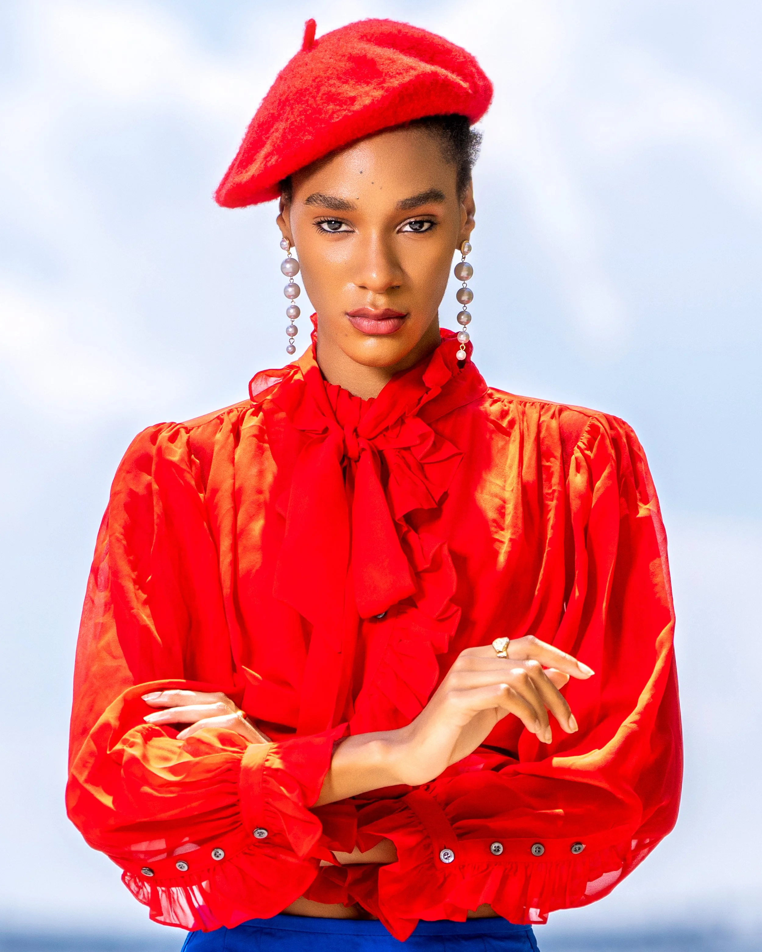 Editorial fashion portrait of a model wearing a vibrant red beret and red ruffled blouse with pearl earrings and a bold blue skirt.
