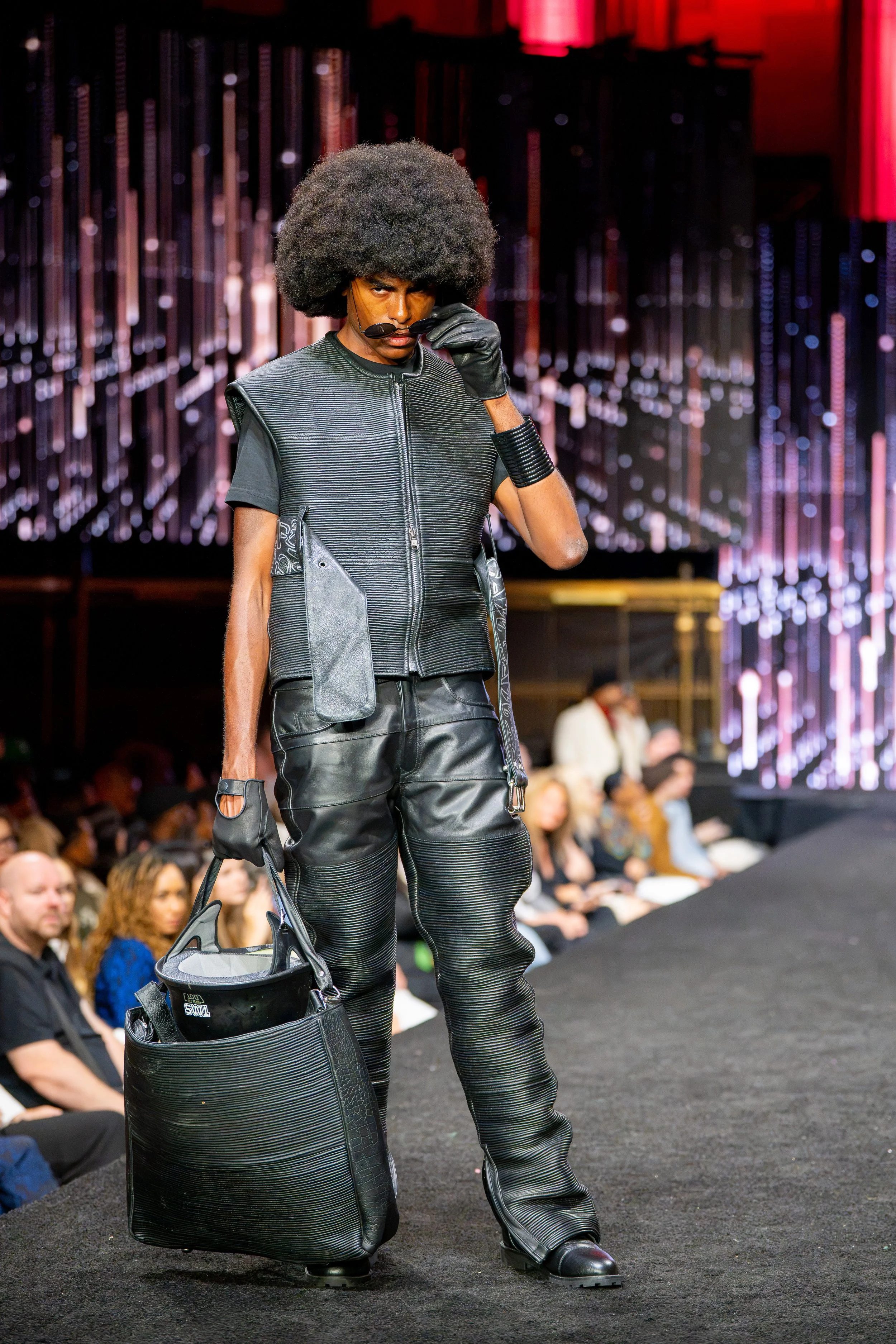Male model with a large afro wearing an all-black leather look and carrying an oversized bag on the New York Fashion Week runway.
