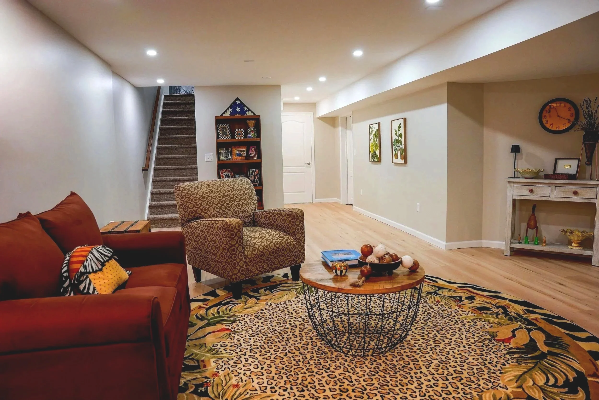 A living room with a brown sofa, a patterned armchair, a round coffee table with decorative balls, and a colorful animal print rug. There are framed pictures on the wall, a tall bookshelf, a clock, and a fireplace with decorations. A staircase leads to an upper level.