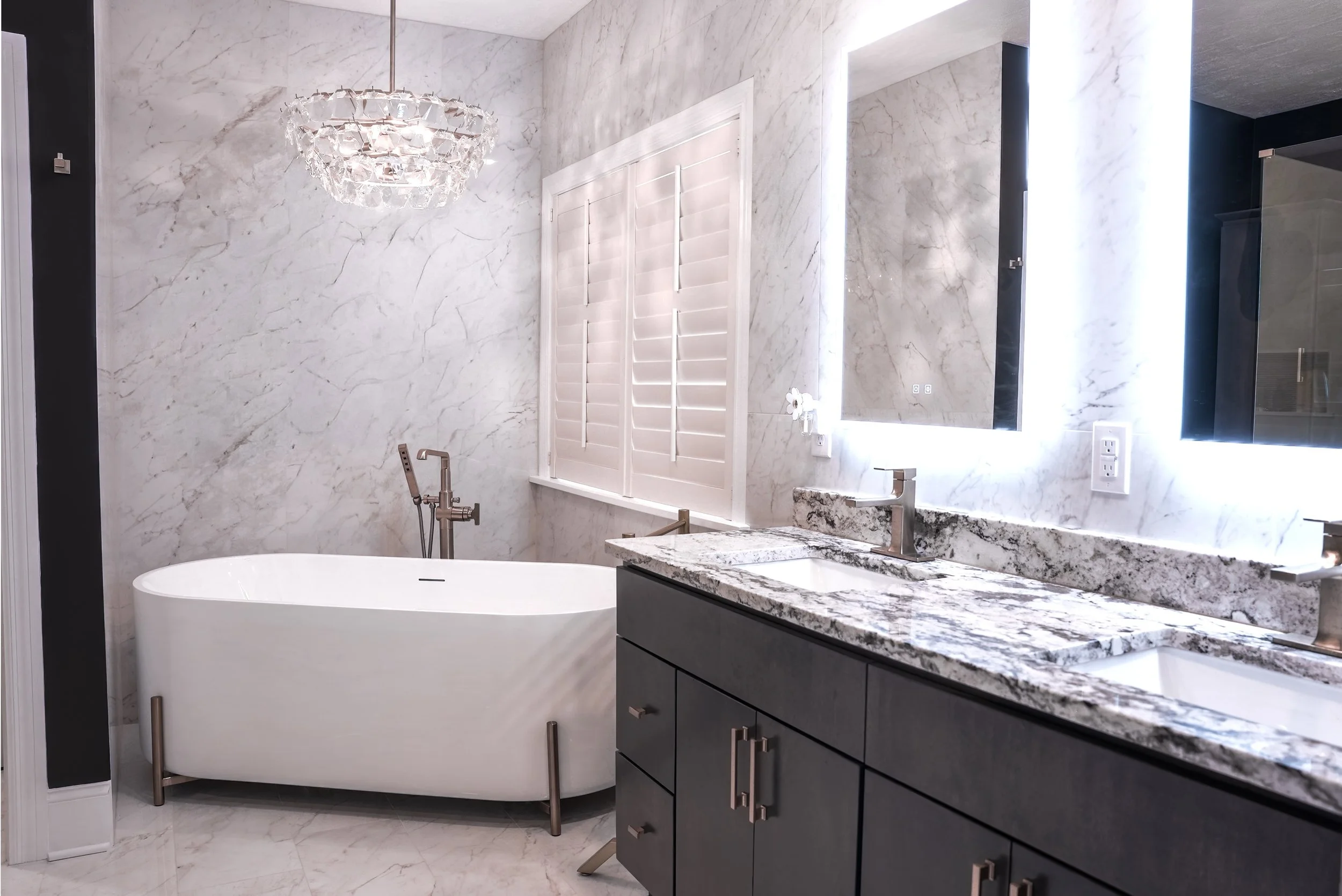 Modern bathroom with marble walls and countertops, a black vanity with silver handles, a white bathtub, a window with white shutters, illuminated mirrors, and a chandelier.