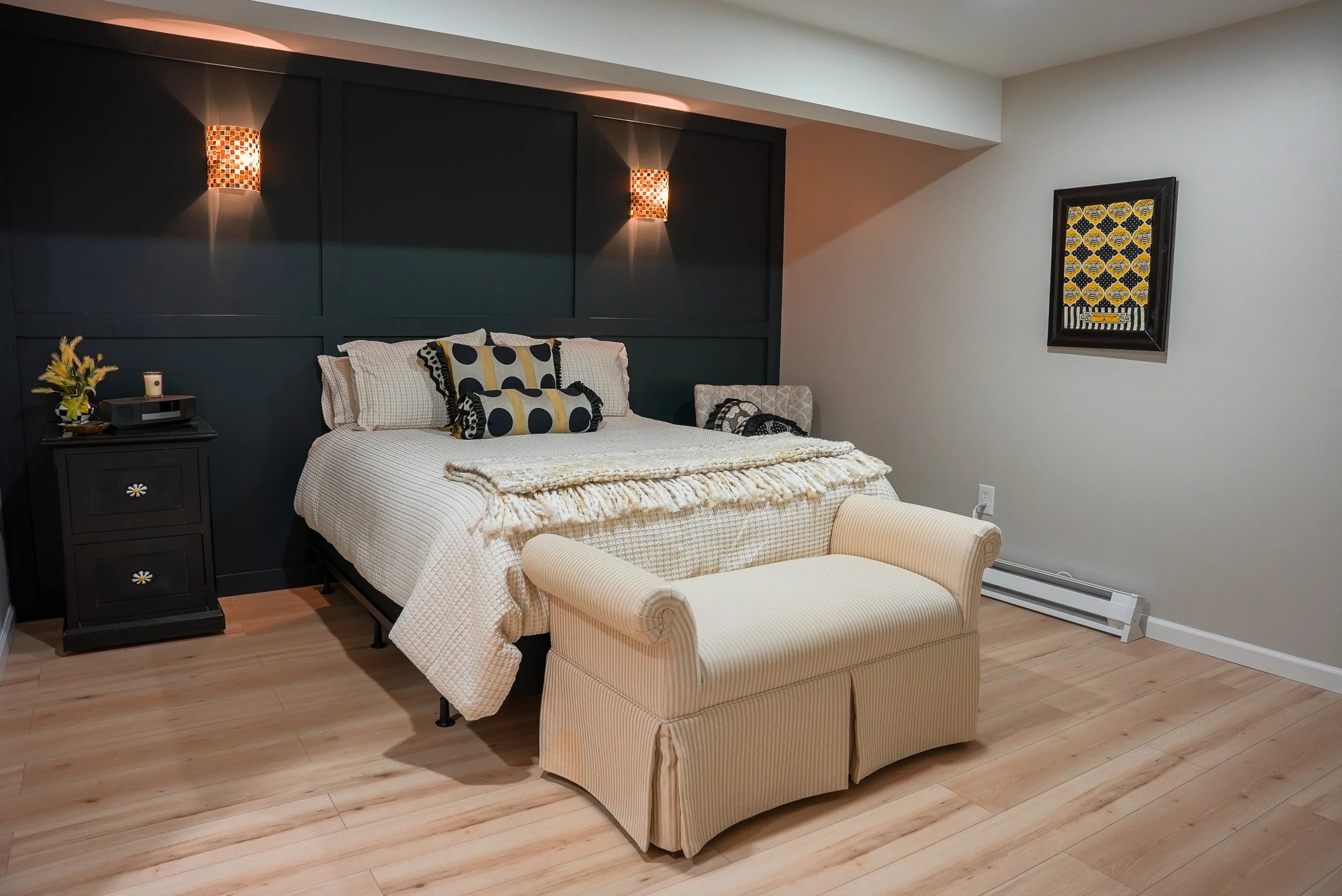 A bedroom with a beige bed, black headboard, and black nightstand. The bed has beige pillows and patterned decorative pillows. There's a beige cushioned bench at the foot of the bed. Two wall-mounted lamps illuminate the headboard. The room has light wooden floors, a framed yellow and black art piece, and white walls with a baseboard heater along the right wall.