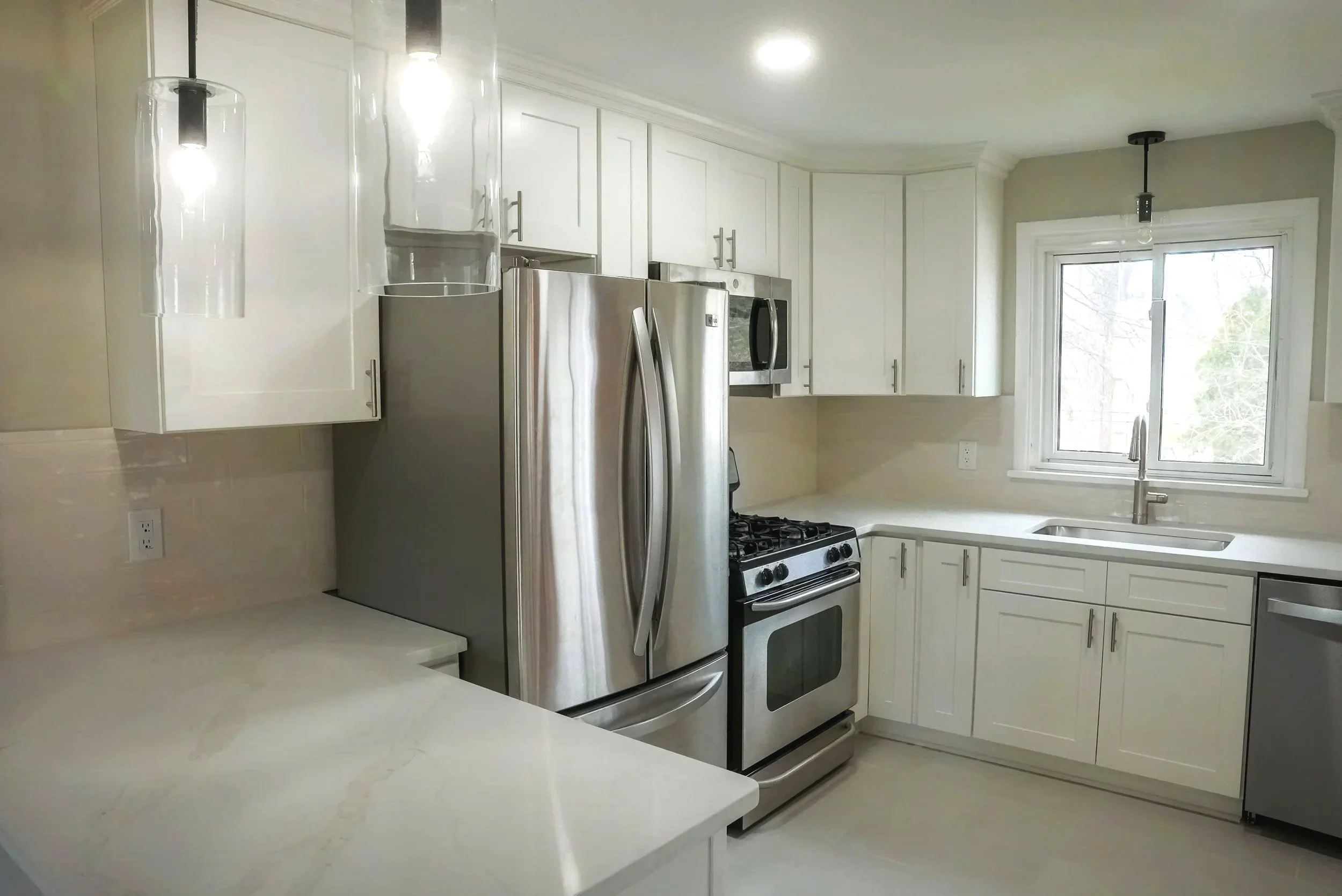 Kitchen with white cabinets, stainless steel refrigerator and oven, white countertops, a window above the sink, and pendant lights.