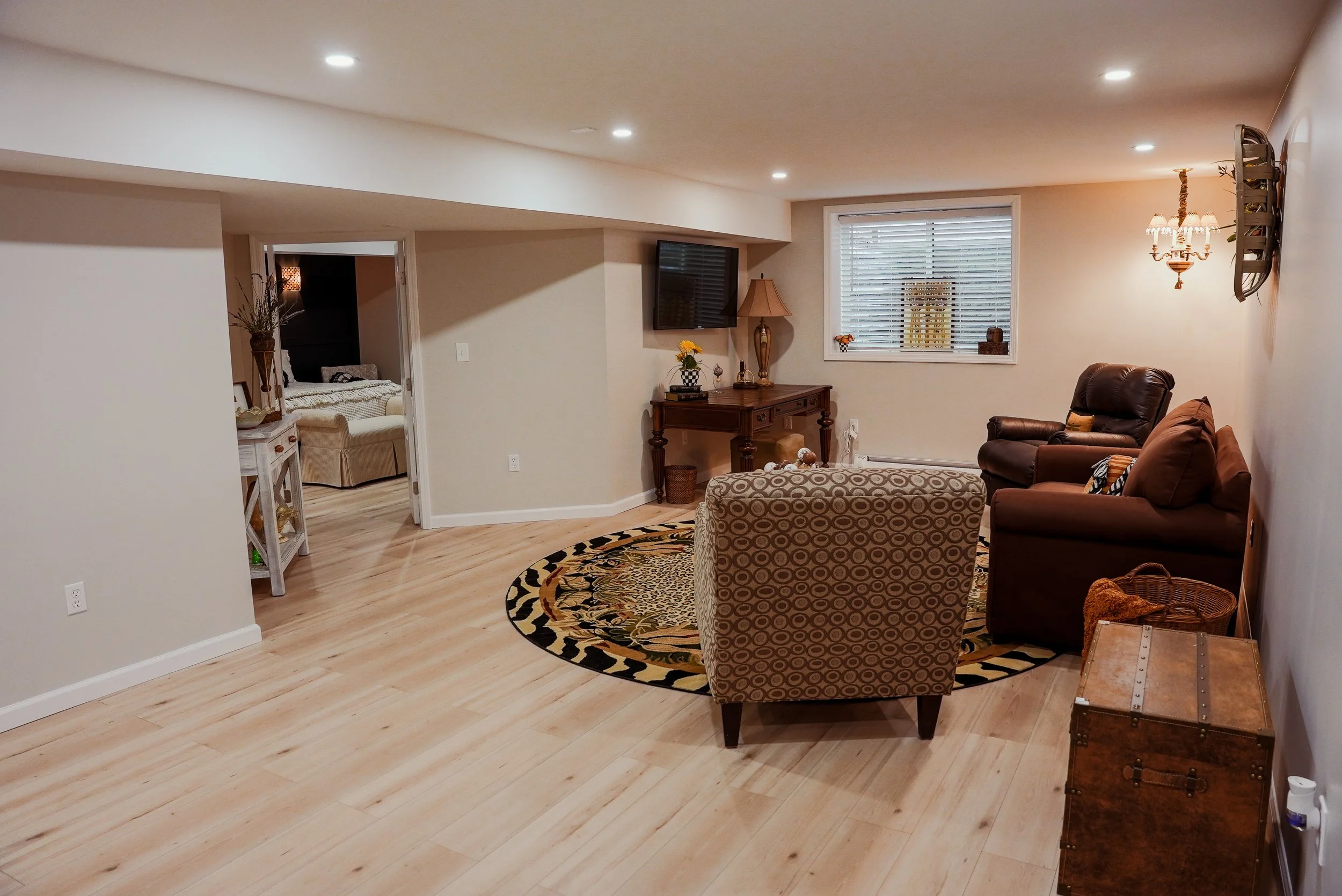 Living room with beige walls, hardwood flooring, a patterned oval rug, a brown sofa, a patterned armchair, a TV mounted on the wall, a wooden console table with a lamp and flowers, and a window with blinds. There is an open doorway leading to a bedroom.