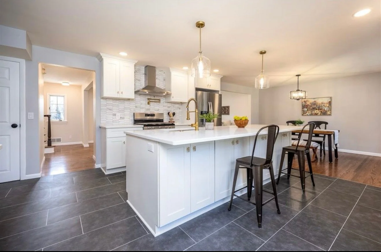 Modern kitchen with white cabinets, a large white island with a gold faucet, black bar stools, stainless steel appliances, pendant lighting, a dining area with a wooden table and black chairs, and hardwood flooring in the dining area.