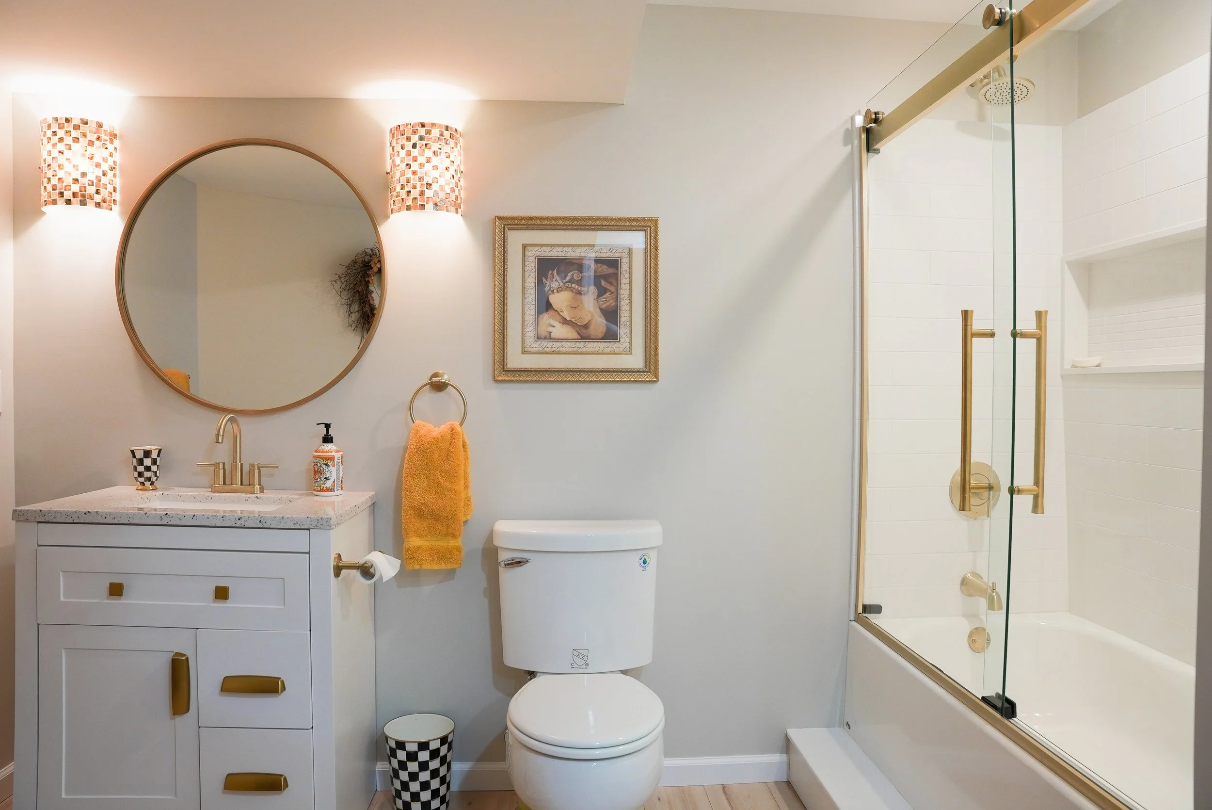 Bathroom with a white vanity, round mirror, framed art, yellow towel, toilet, checkered trash bin, and glass shower with brass fixtures.