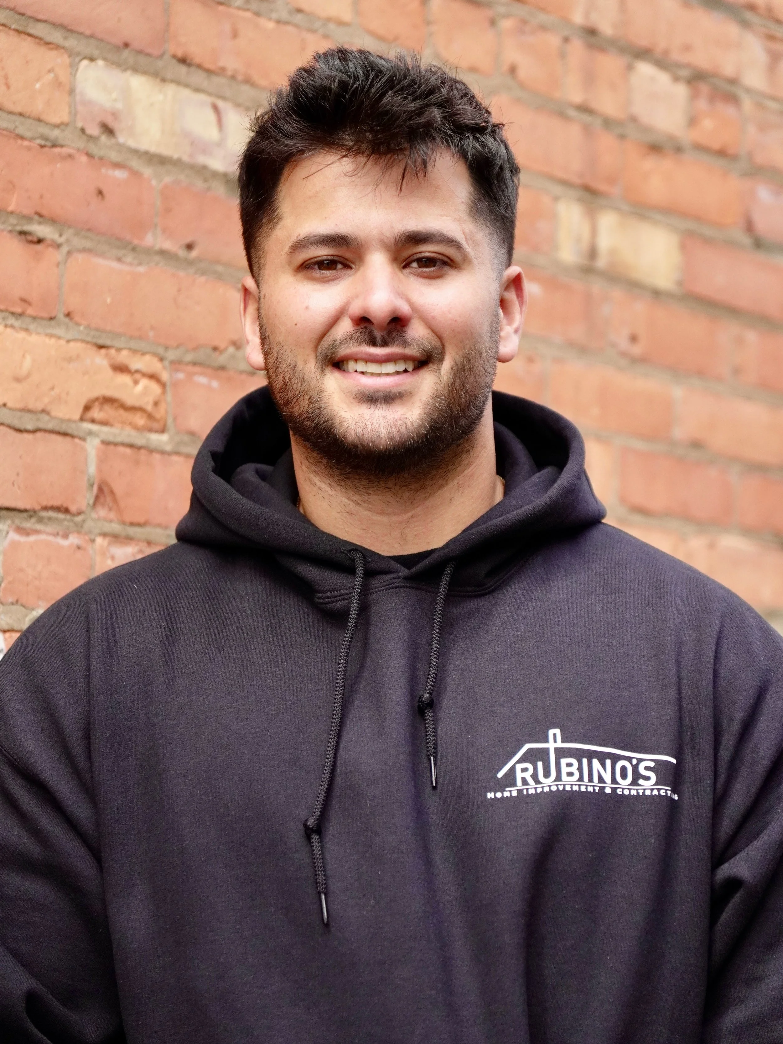 Close-up of a young man with dark hair, facial hair, and light skin, wearing a black hoodie with a 'Rubino's Home Improvement & Contracting' logo, standing against a brick wall.