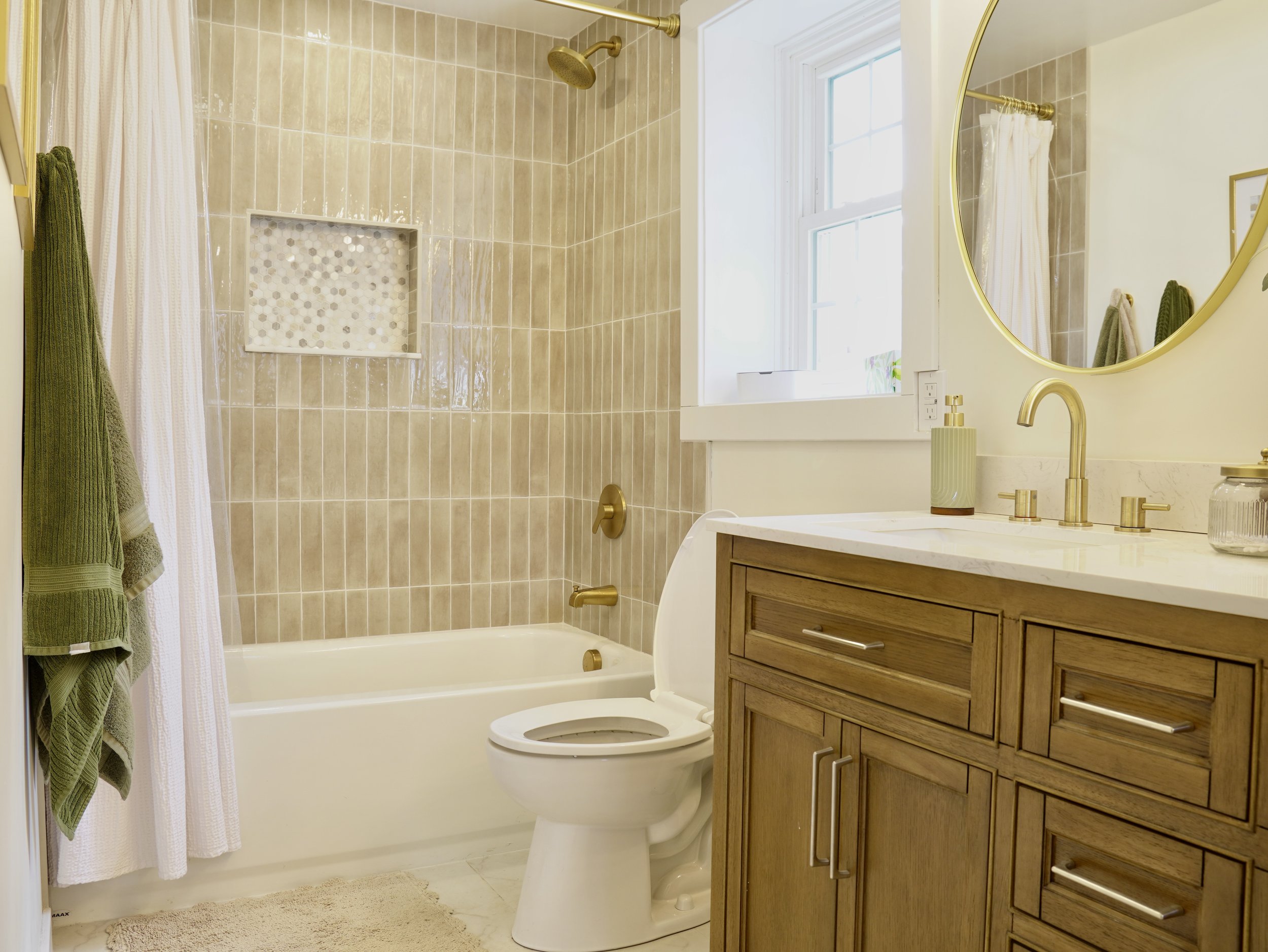 A bathroom with a bathtub, toilet, wooden vanity with a marble countertop, round mirror, gold fixtures, a window, and green towels hanging on a rack.