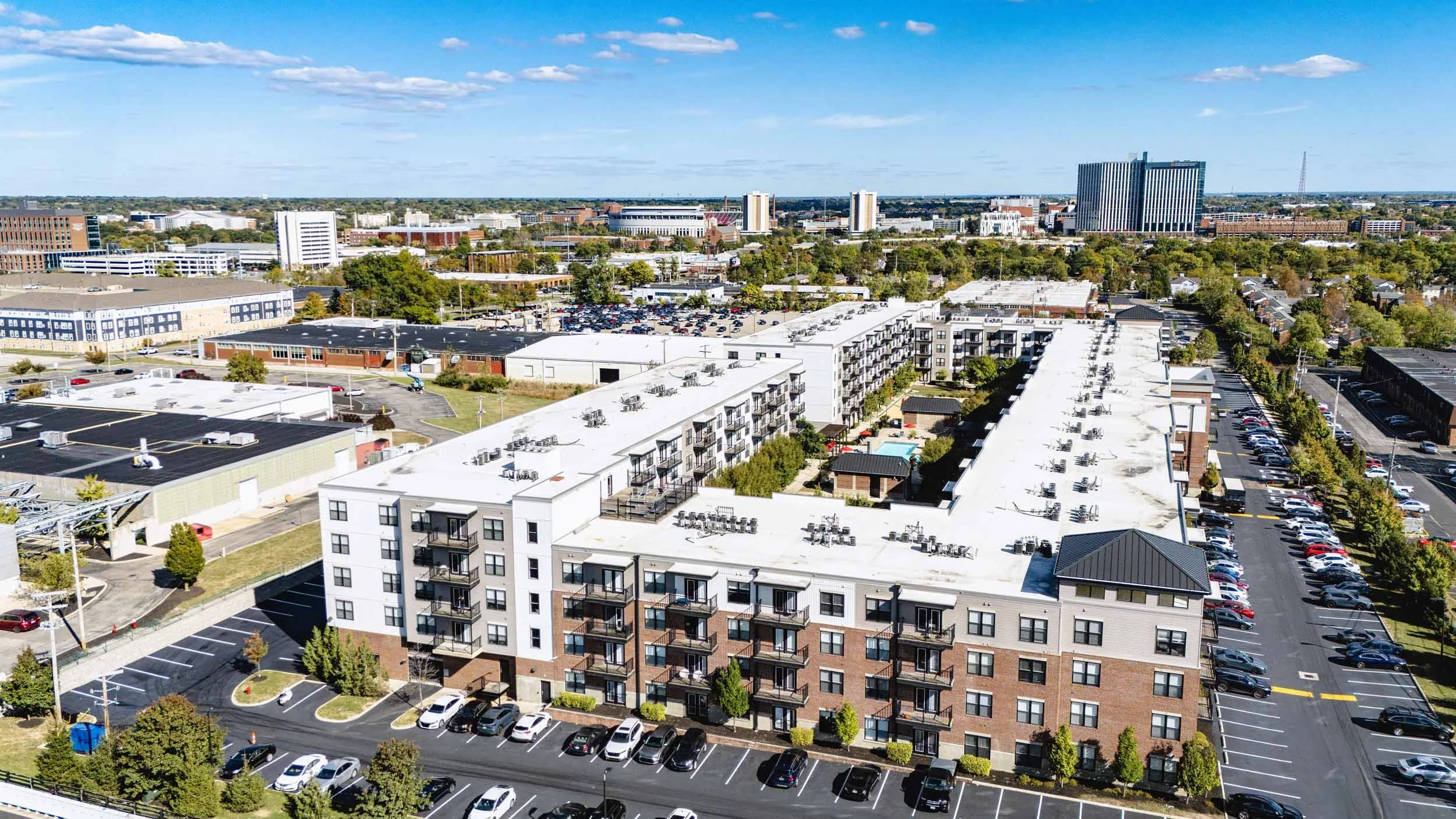 Aerial view of a cityscape showing a multi-story apartment complex with parking lots, surrounding commercial buildings, and city buildings in the background under a partly cloudy sky.