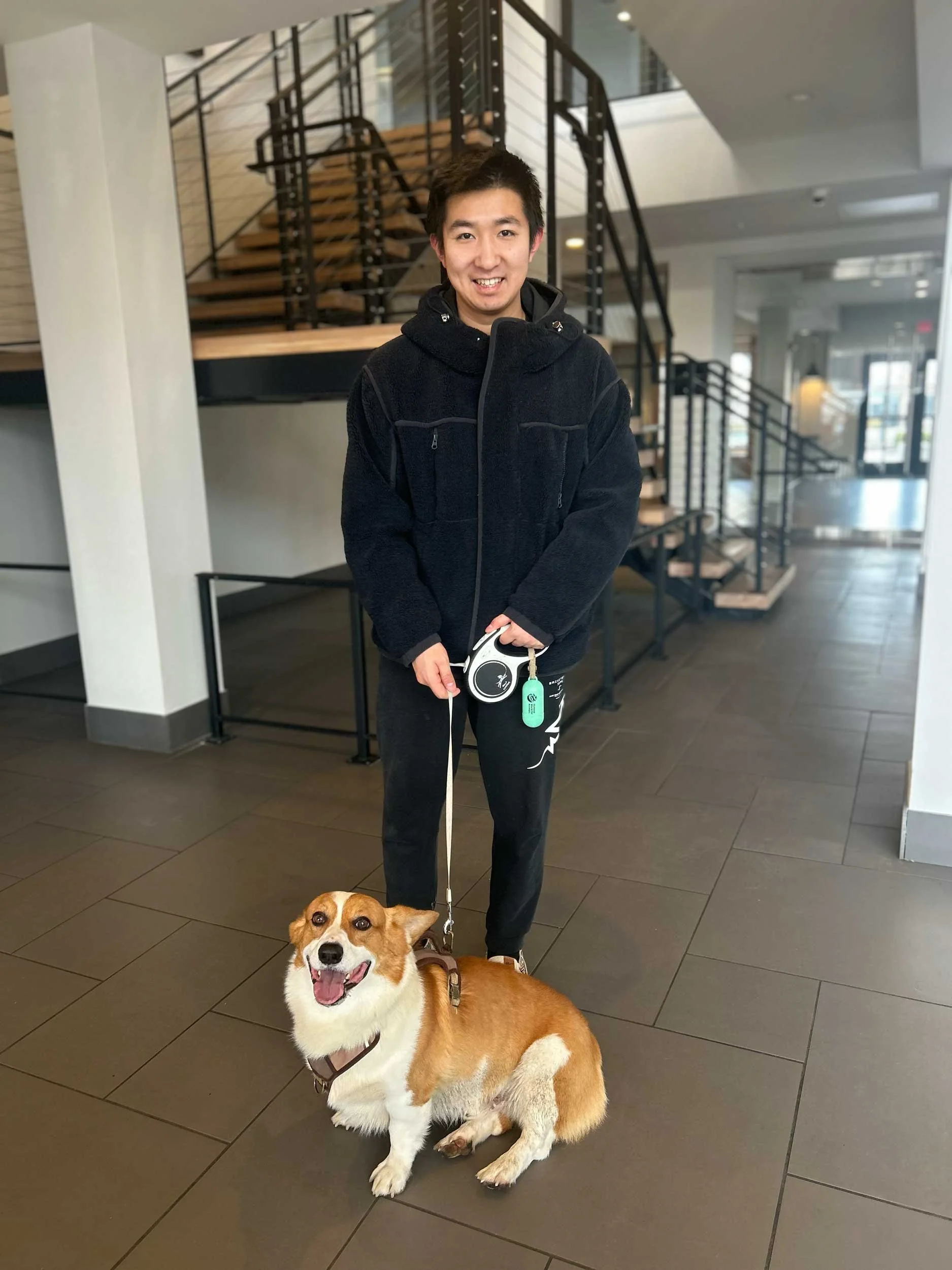 A young man smiling and holding a leash attached to a happy, sitting corgi in an indoor setting with stairs in the background.