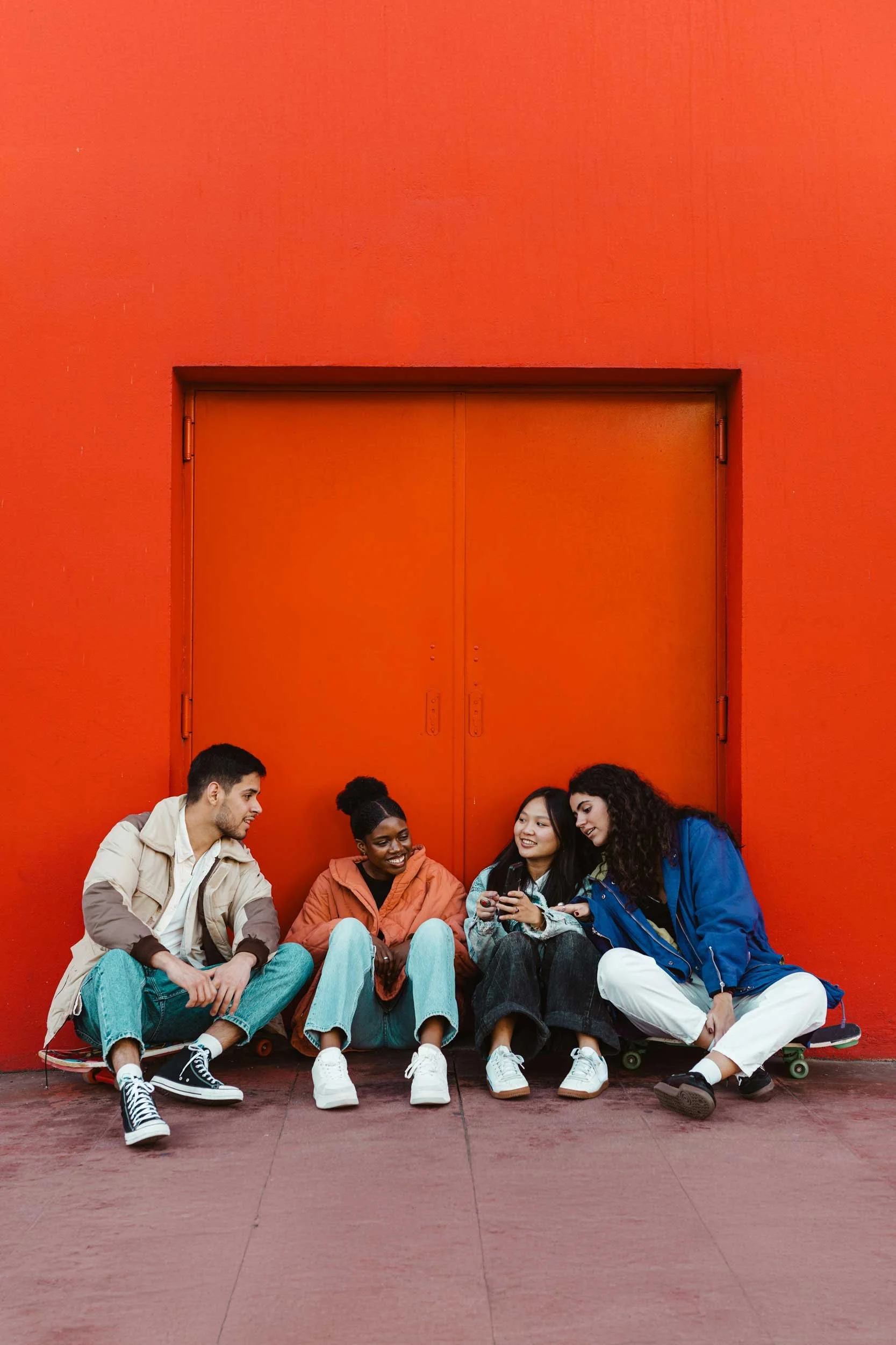 Four friends sitting on the ground, smiling and looking at a phone, against a bright red wall, with skateboards.