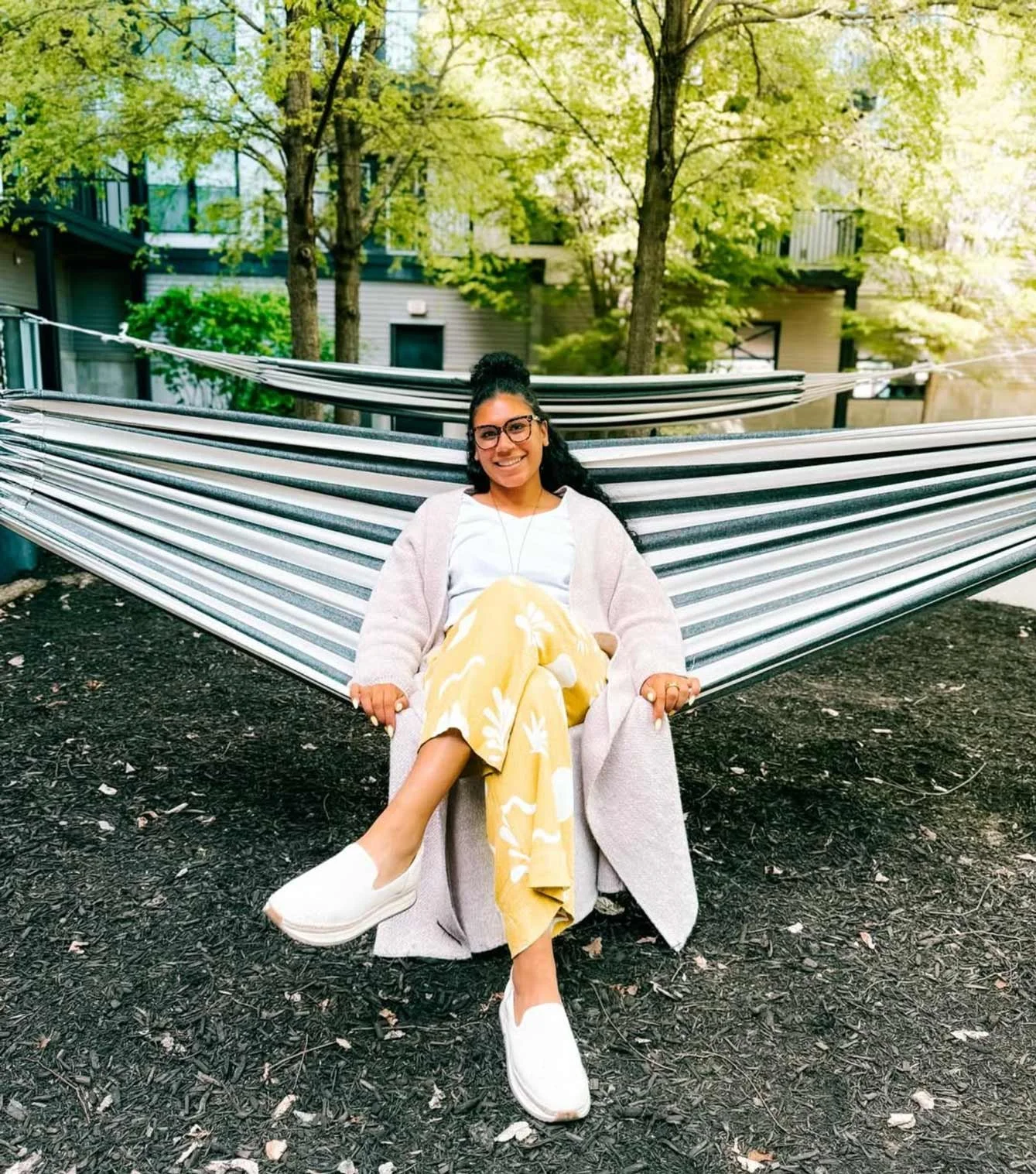 A woman with glasses and dark hair tied back, smiling, sitting on a hammock outdoors with trees and buildings in the background.
