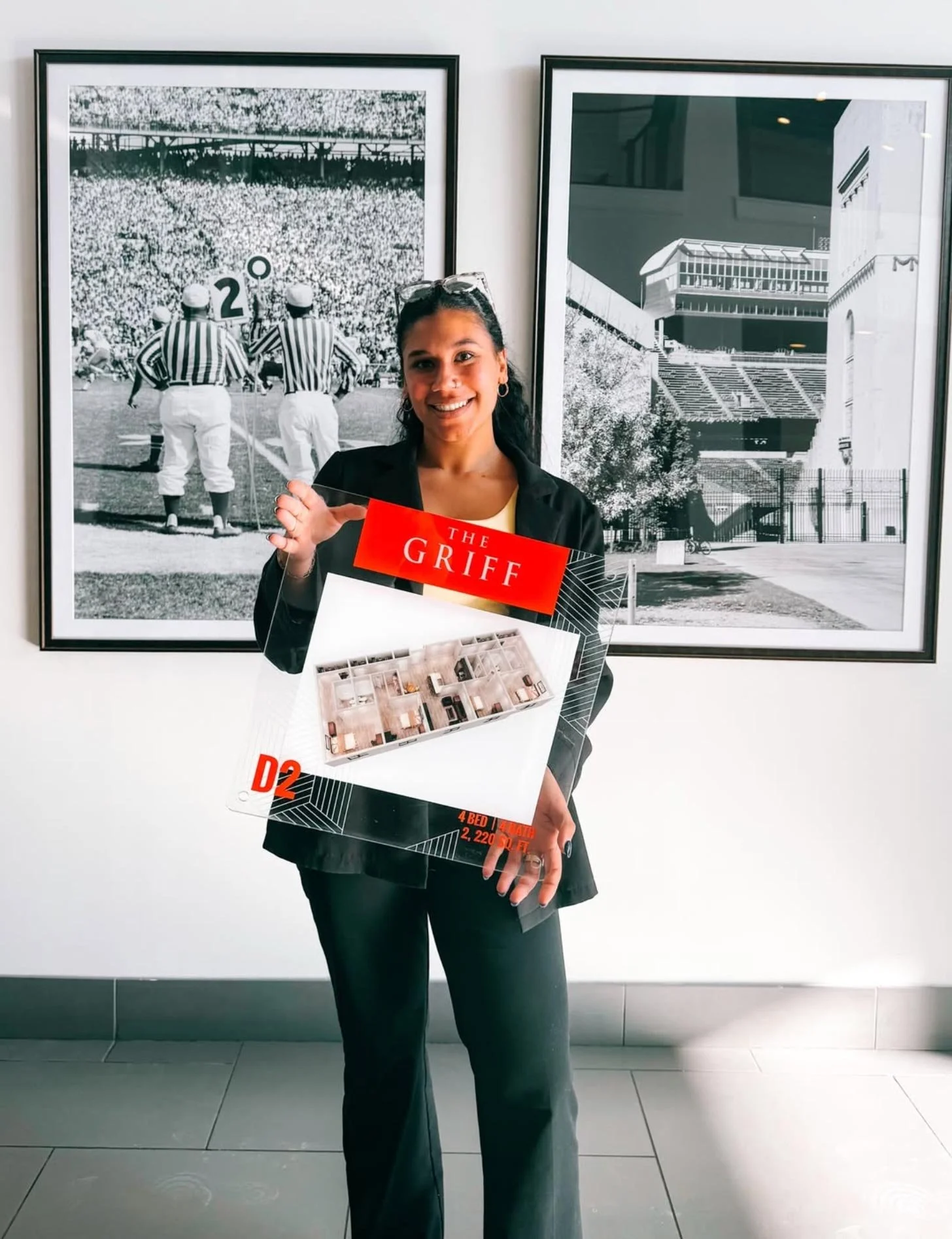 A woman with dark hair, smiling, wearing a black blazer, holding a real estate brochure, standing in front of black and white photographs of stadiums and sports events.