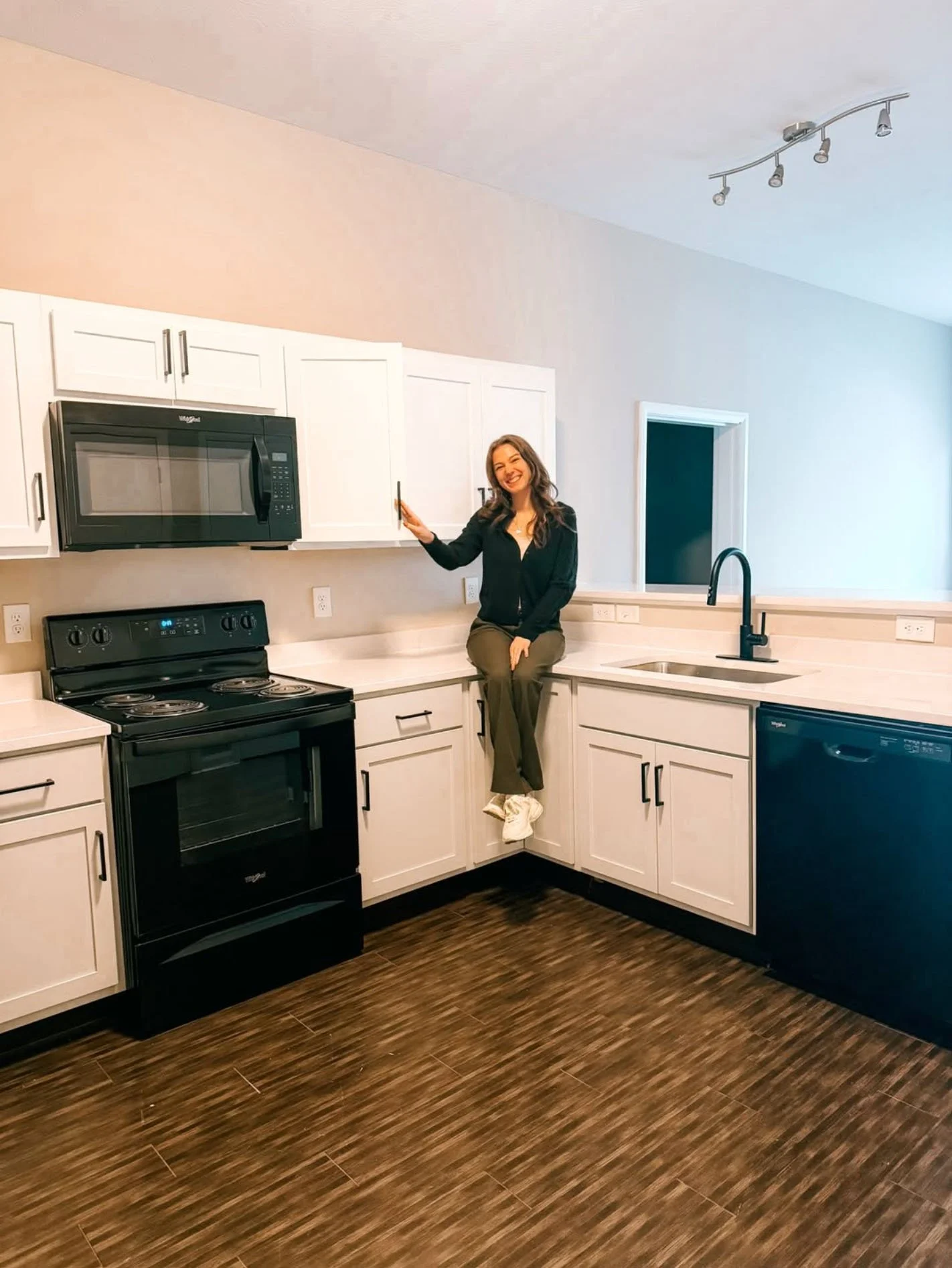 A woman sitting on a kitchen counter with white cabinets, a black stove, a microwave, a black dishwasher, and a black faucet in a modern kitchen.