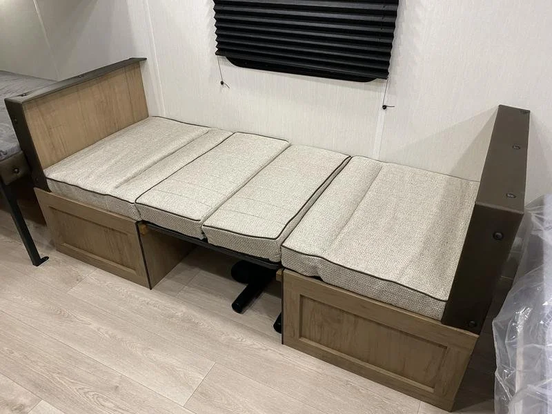 A wooden daybed with a fold-out trundle bed, with beige cushions on top and a black window blind on the wall behind it.