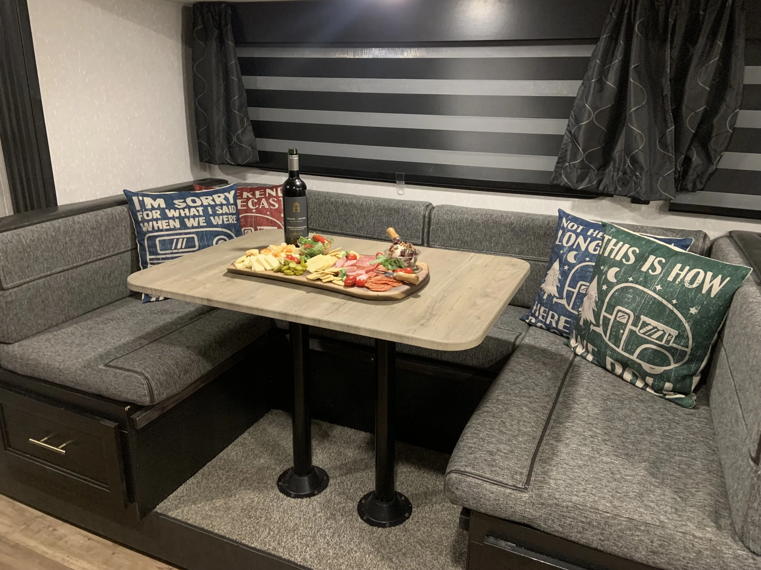 A U-shaped gray sofa with colorful throw pillows, a wooden table with cheese, meat, crackers, and a bottle of wine in a cozy room with dark striped window shades and black curtains.