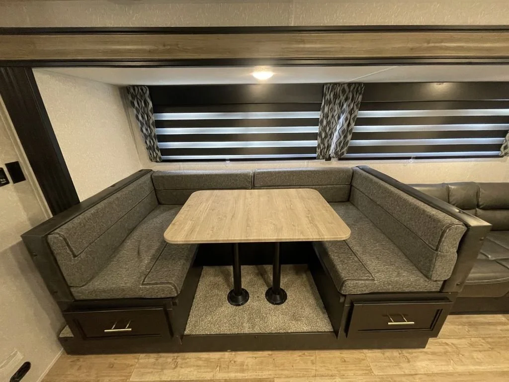A built-in corner dining nook with gray upholstered cushions and a wooden table, situated in a room with patterned curtains and blinds on the windows.